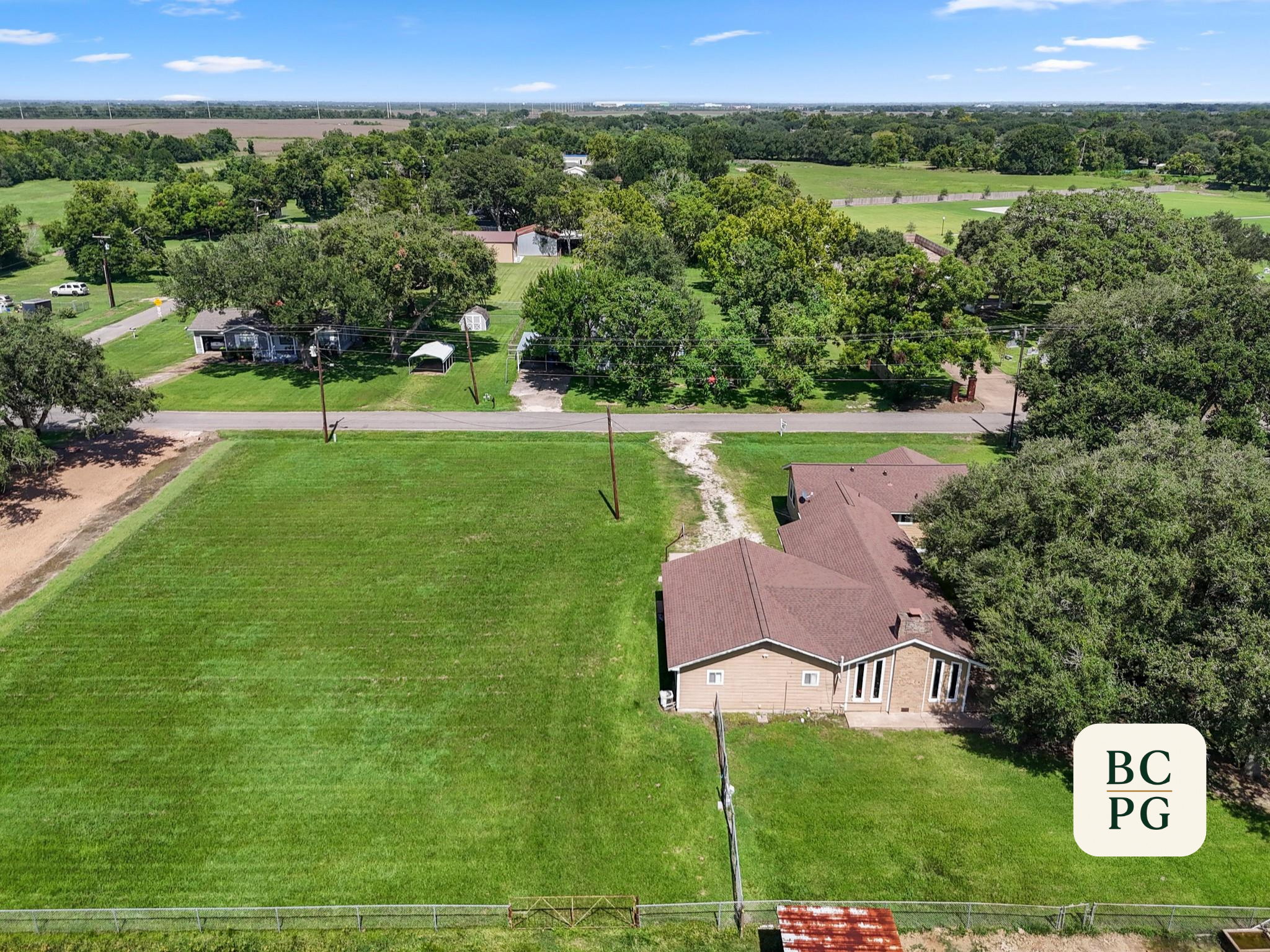 An aerial view of a suburban neighborhood with houses, trees, and grassy yards, with open fields and a blue sky in the background.