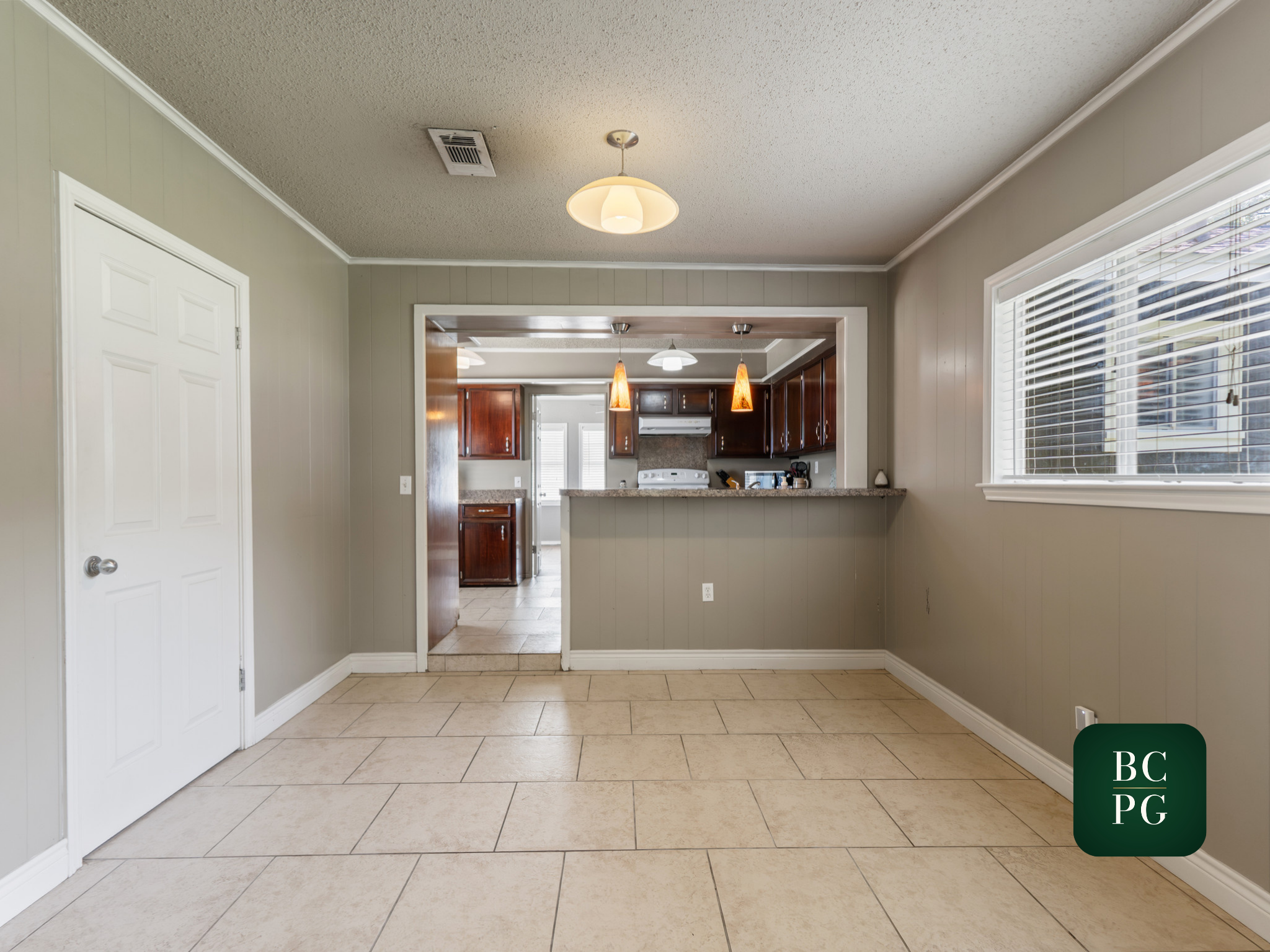 An interior view of a home showing a tiled floor, beige walls, a ceiling with a light fixture, and a kitchen with dark wood cabinets and white appliances visible through an open doorway.