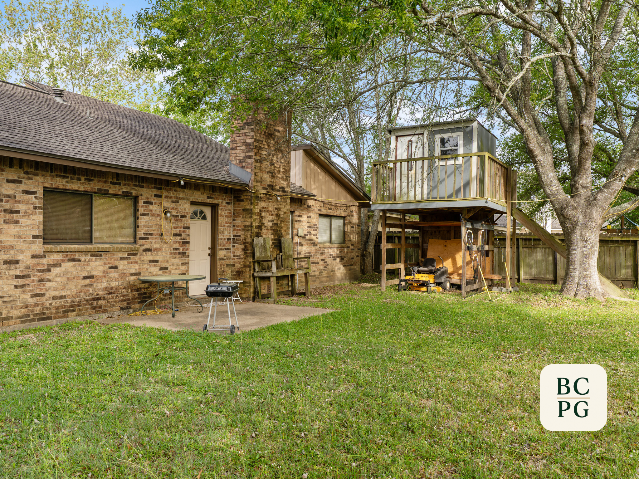 Backyard scene with brick house, large trees, a small porch area, lawn, and various outdoor equipment like a lawn mower, table, and ladder.