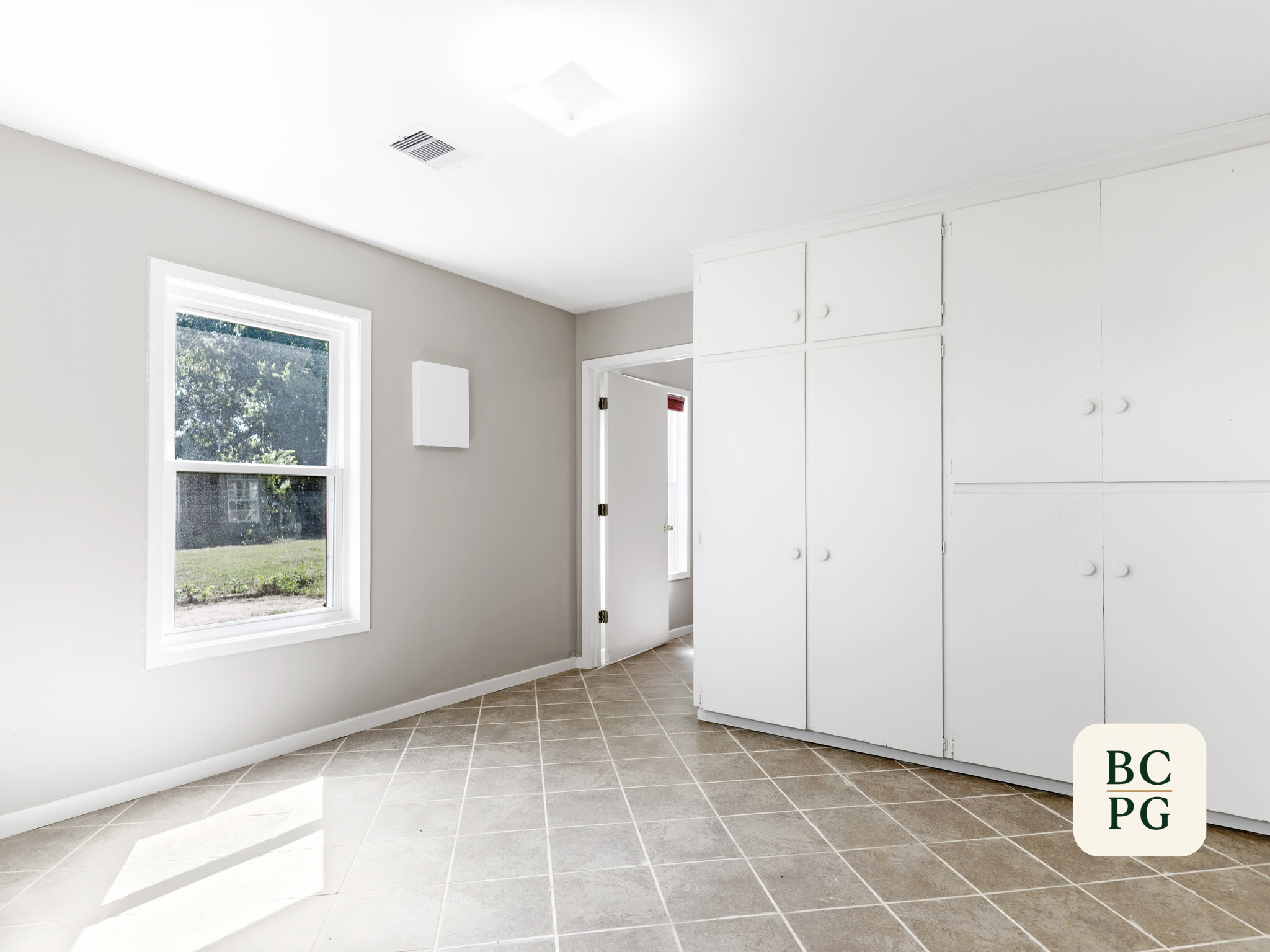 A bright white kitchen with tile flooring, a window revealing a green lawn outside, and white cabinets with round knobs.