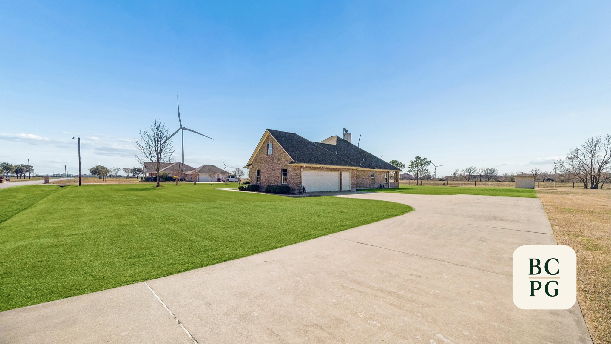 A suburban house with a two-car garage, surrounded by a well-maintained lawn, in a rural area with wind turbines in the background, under a clear blue sky.