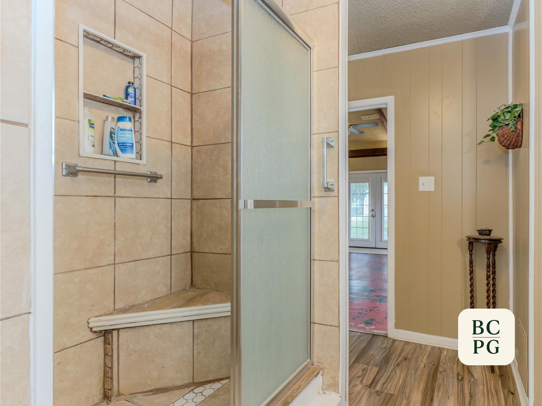 Bathroom with a beige tiled shower, a built-in shelf with toiletries, a grab bar, a frosted glass shower door, and a view into another room with a window and wooden floors.
