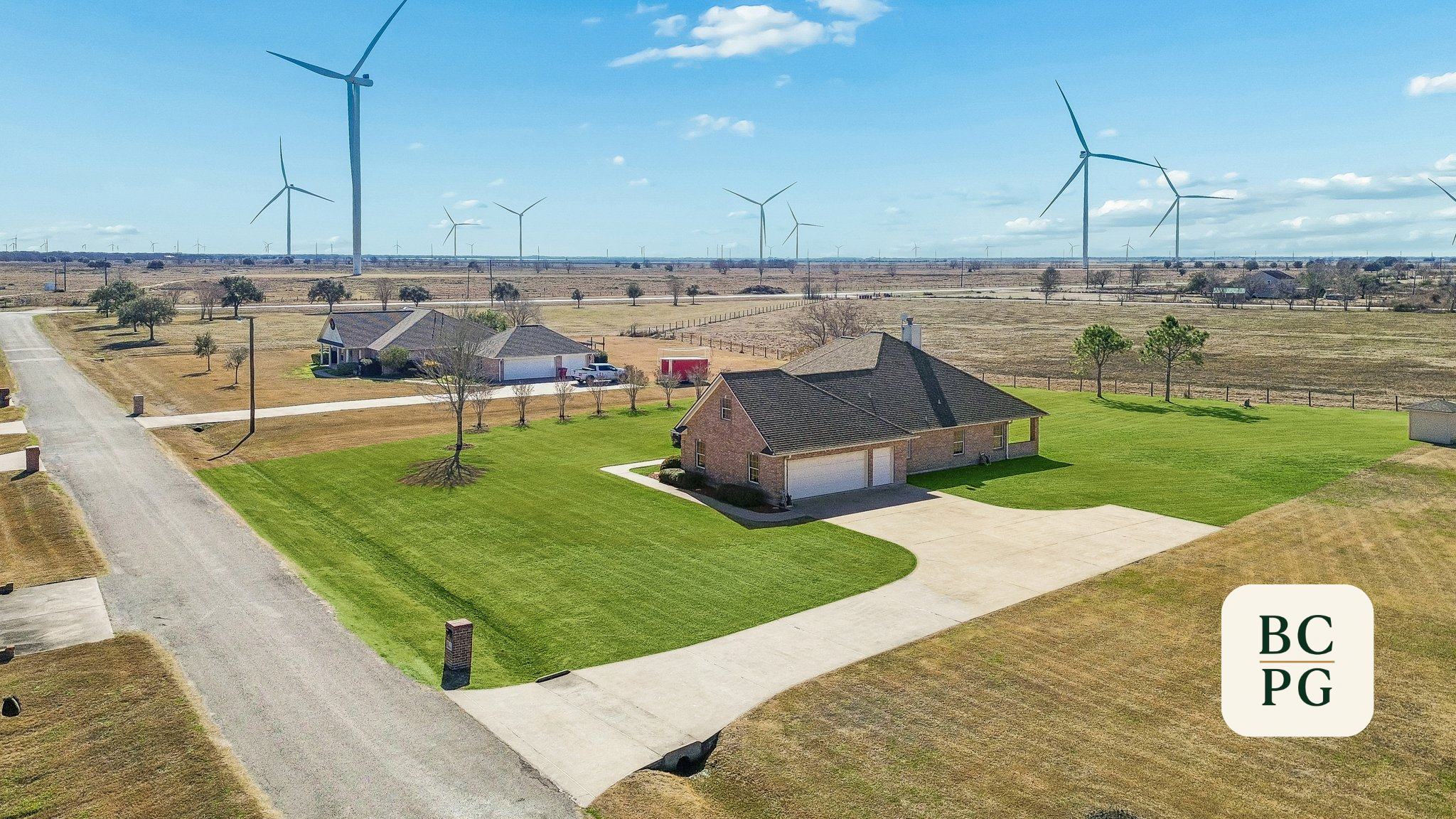 Aerial view of a suburban house with a green lawn, trees, driveway, and neighboring houses, with wind turbines in the distance under a blue sky with scattered clouds.