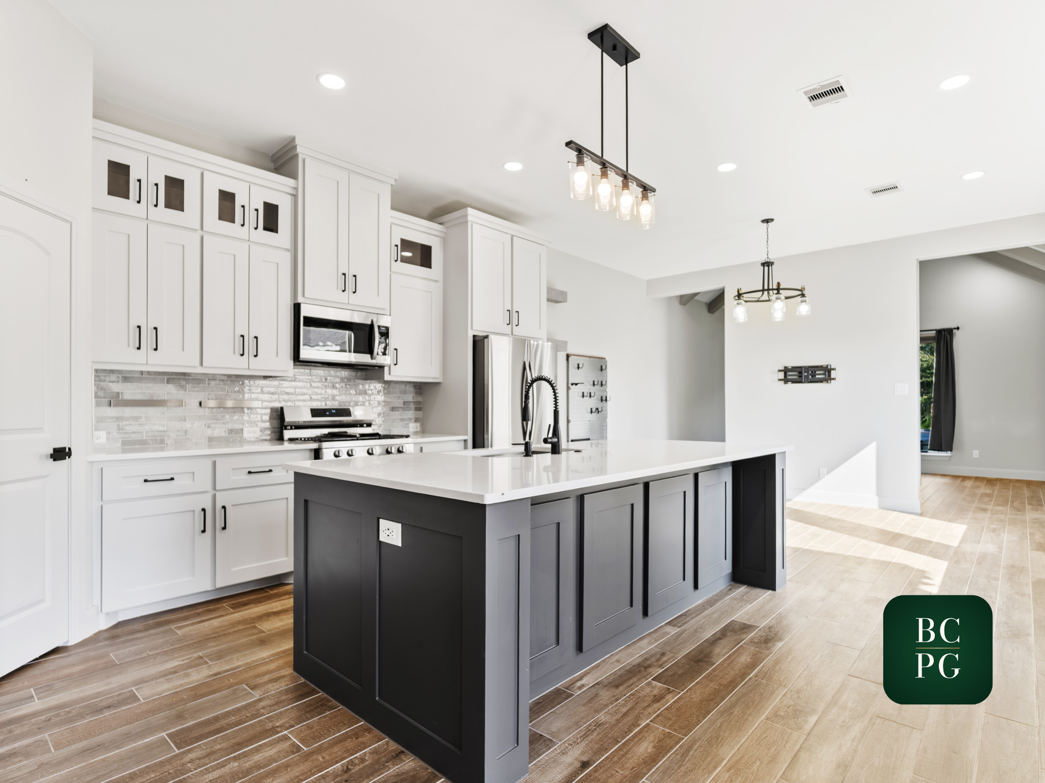 Modern kitchen with white cabinets, stainless steel appliances, a dark gray kitchen island, wood flooring, and black pendant and ceiling lights.