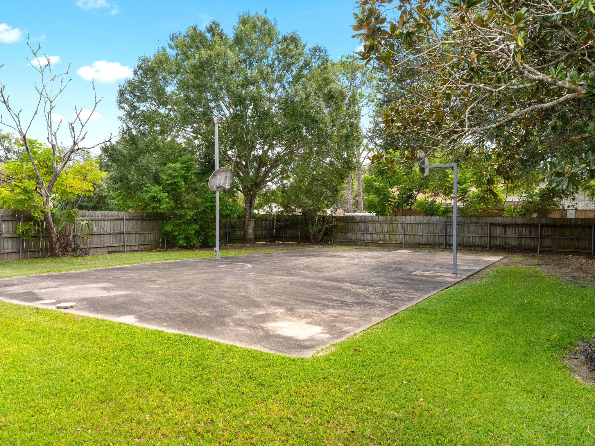 An outdoor basketball court with two hoops, surrounded by green grass and trees, enclosed by a wooden fence.
