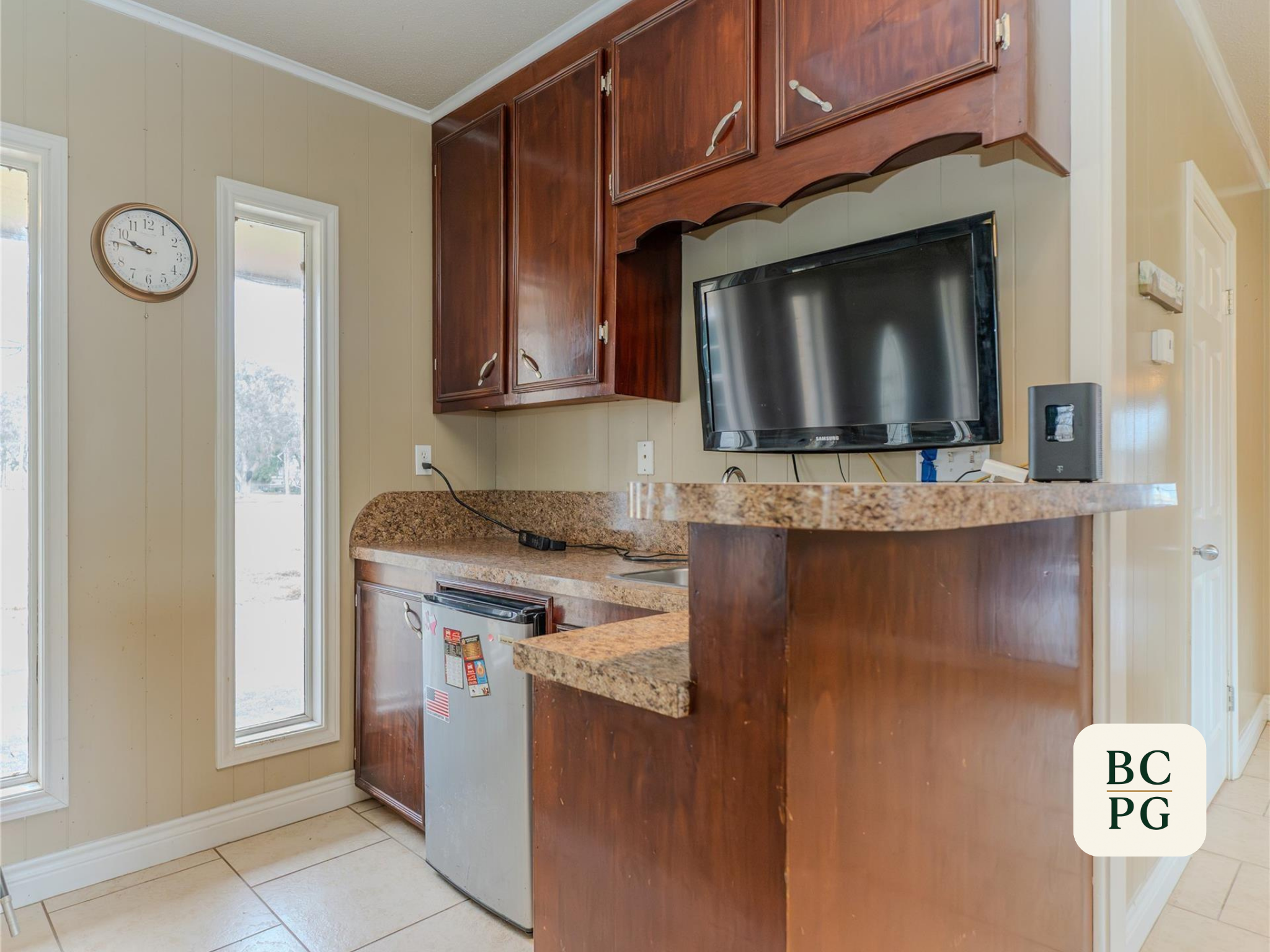 View of a kitchen corner with beige walls, a wall clock, two windows, and cherry wood cabinets. A television is mounted above a granite countertop, with a small refrigerator below.