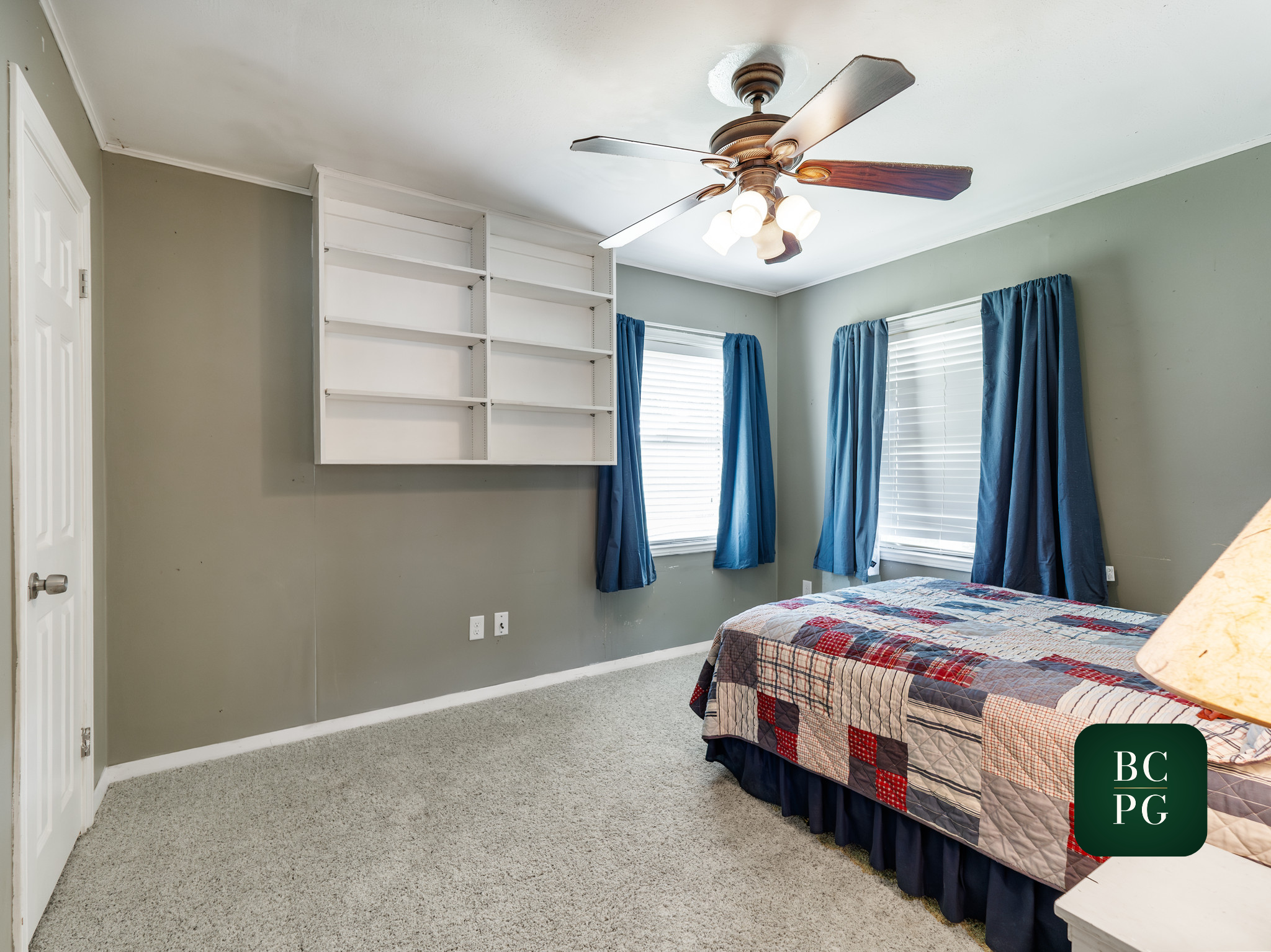 A bedroom with gray walls, a ceiling fan with wooden blades and lights, two windows with white blinds and blue curtains, and a bed with a checkered quilt.