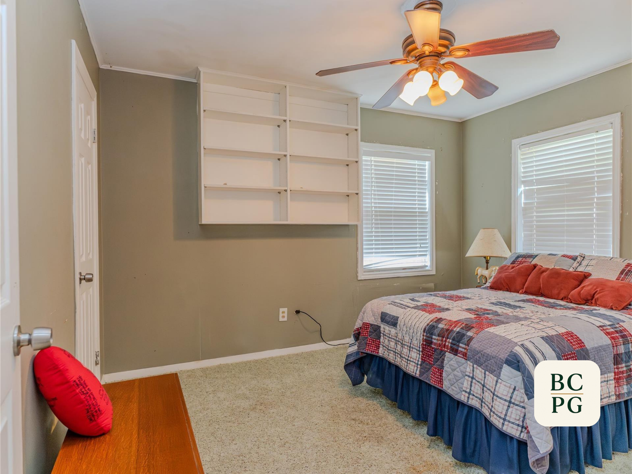 A bedroom with green walls, beige carpet, and a bed with a plaid quilt. Two windows with white blinds and a wooden ceiling fan with lights. A bookshelf is built into the wall, and a red pillow is on a wooden bench near the door.