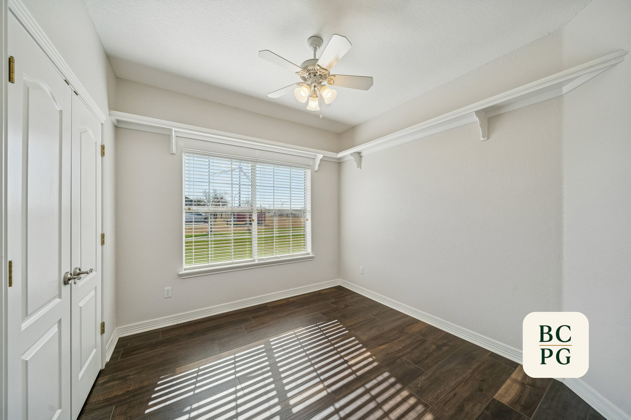 Empty room with white walls, dark wood flooring, a window with blinds, a ceiling fan with lights, and double closet doors.