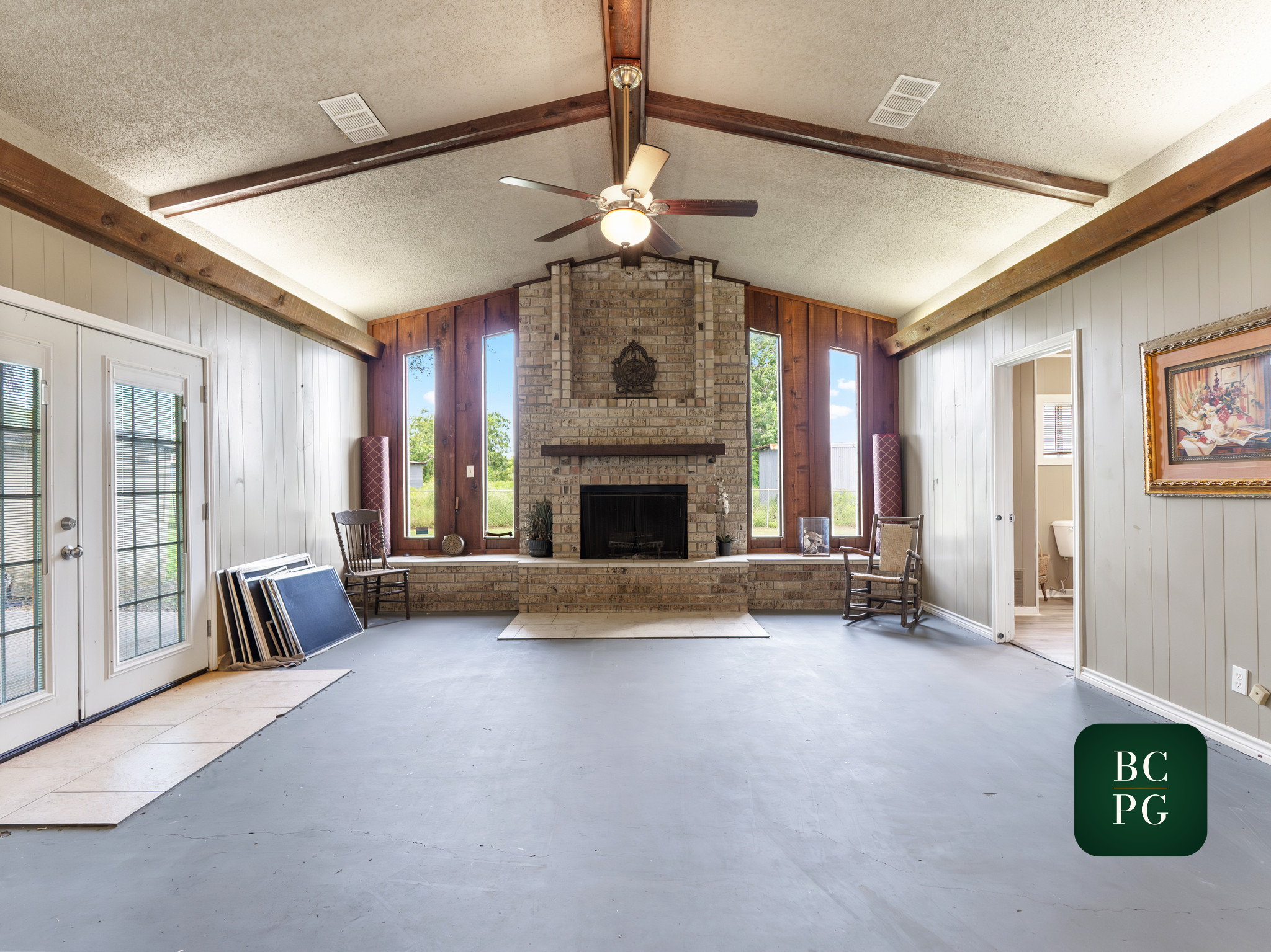 Interior room with a brick fireplace, wooden beams on the ceiling, and large windows letting in natural light. There are chairs, a picture on the wall, and construction materials on the floor.