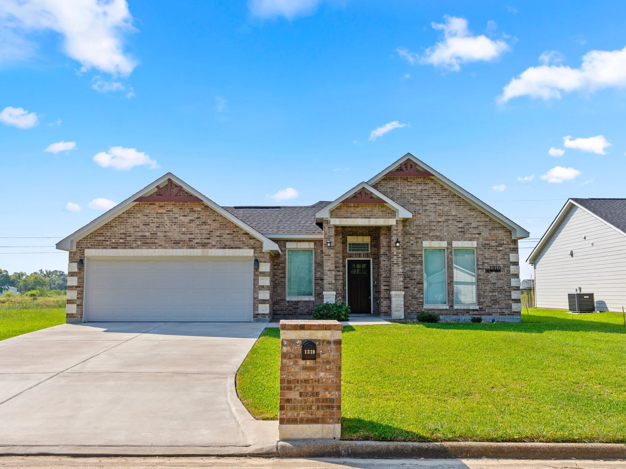 Front view of a brick house with a manicured lawn, driveway, and a mailbox with the house number 1310, under a bright blue sky with scattered clouds.