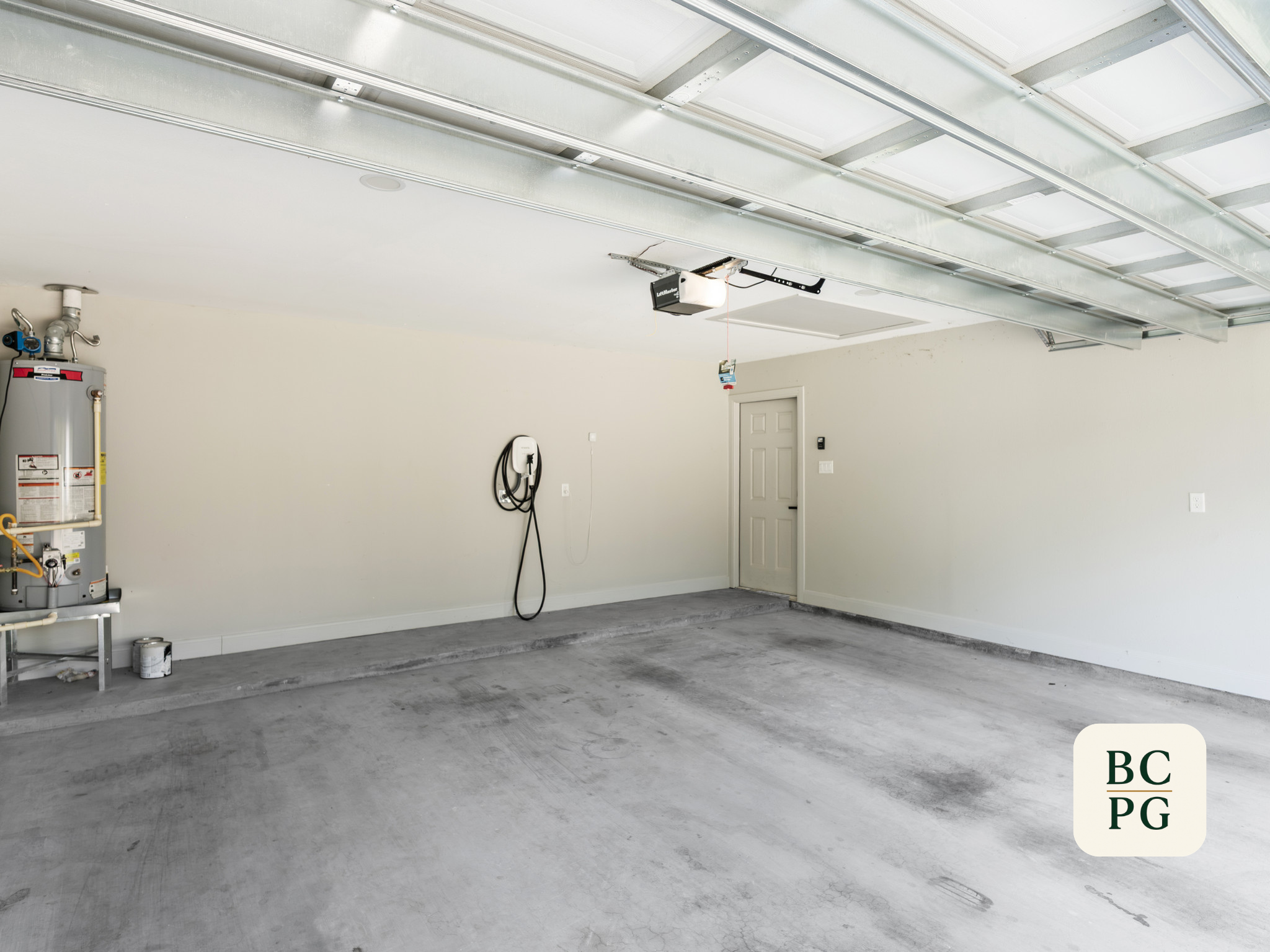 Empty garage with a concrete floor, a water heater on the left, and a closed door in the back. The garage door is partially open, revealing the garage opener on the ceiling side.