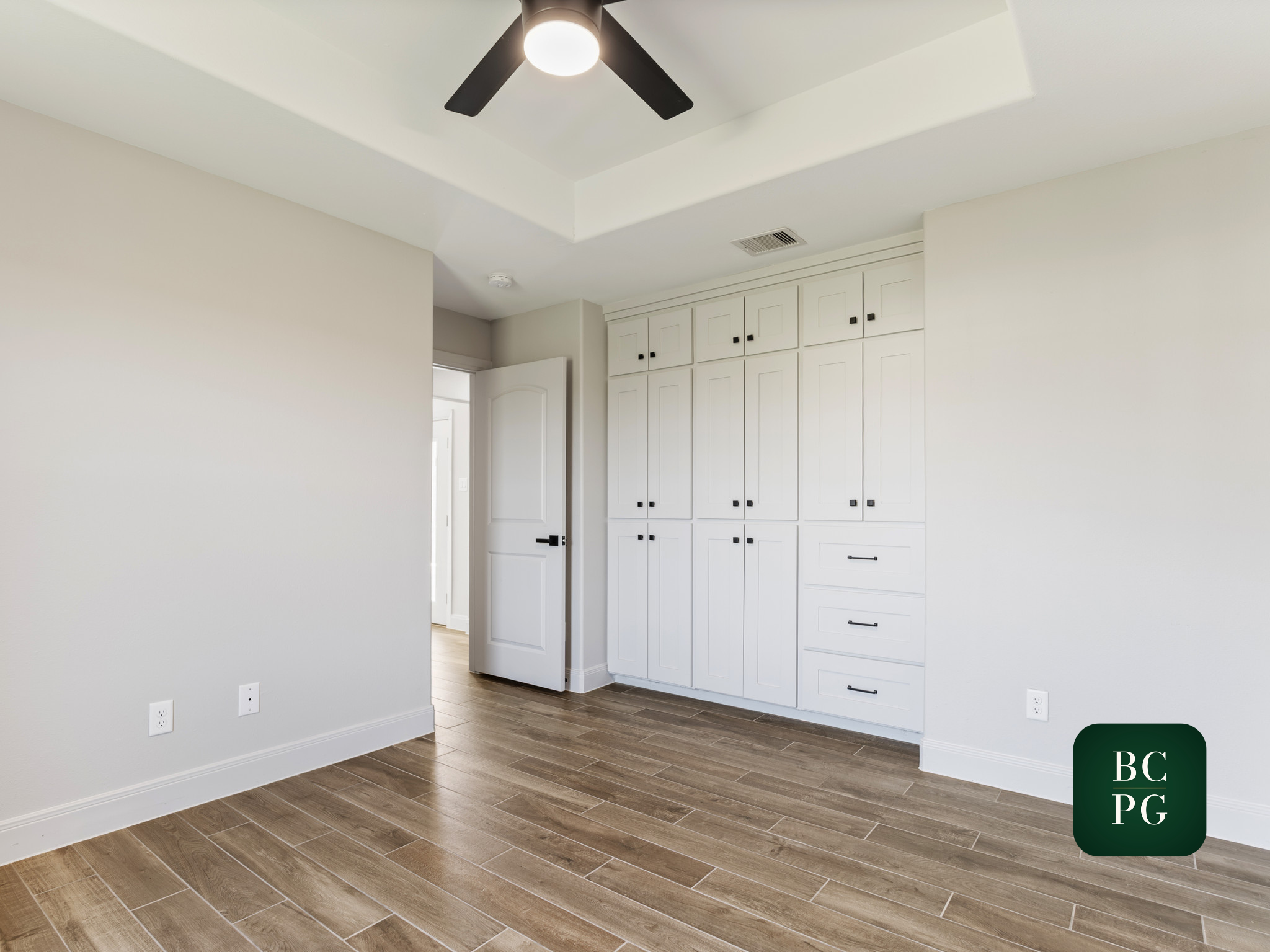 A room with white walls, a ceiling fan with lights, a door slightly open, and built-in white cabinets with black handles. The floor has wood-look tiles.