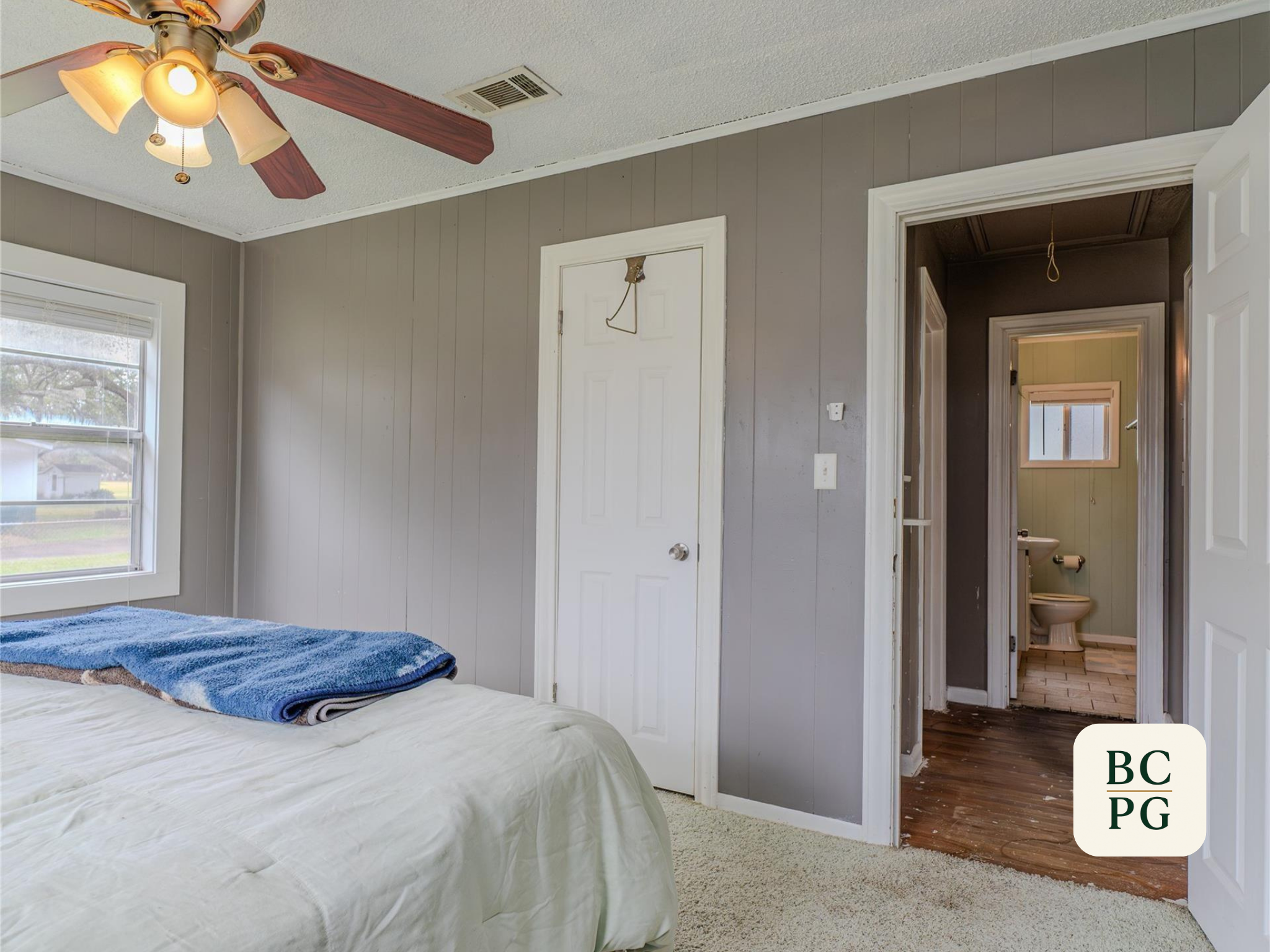 Interior of a bedroom with a bed in the foreground, a window on the left, and an open door leading to a bathroom. The walls are painted gray, and there is a ceiling fan with lights. The bathroom appears to have a separate toilet area visible through 