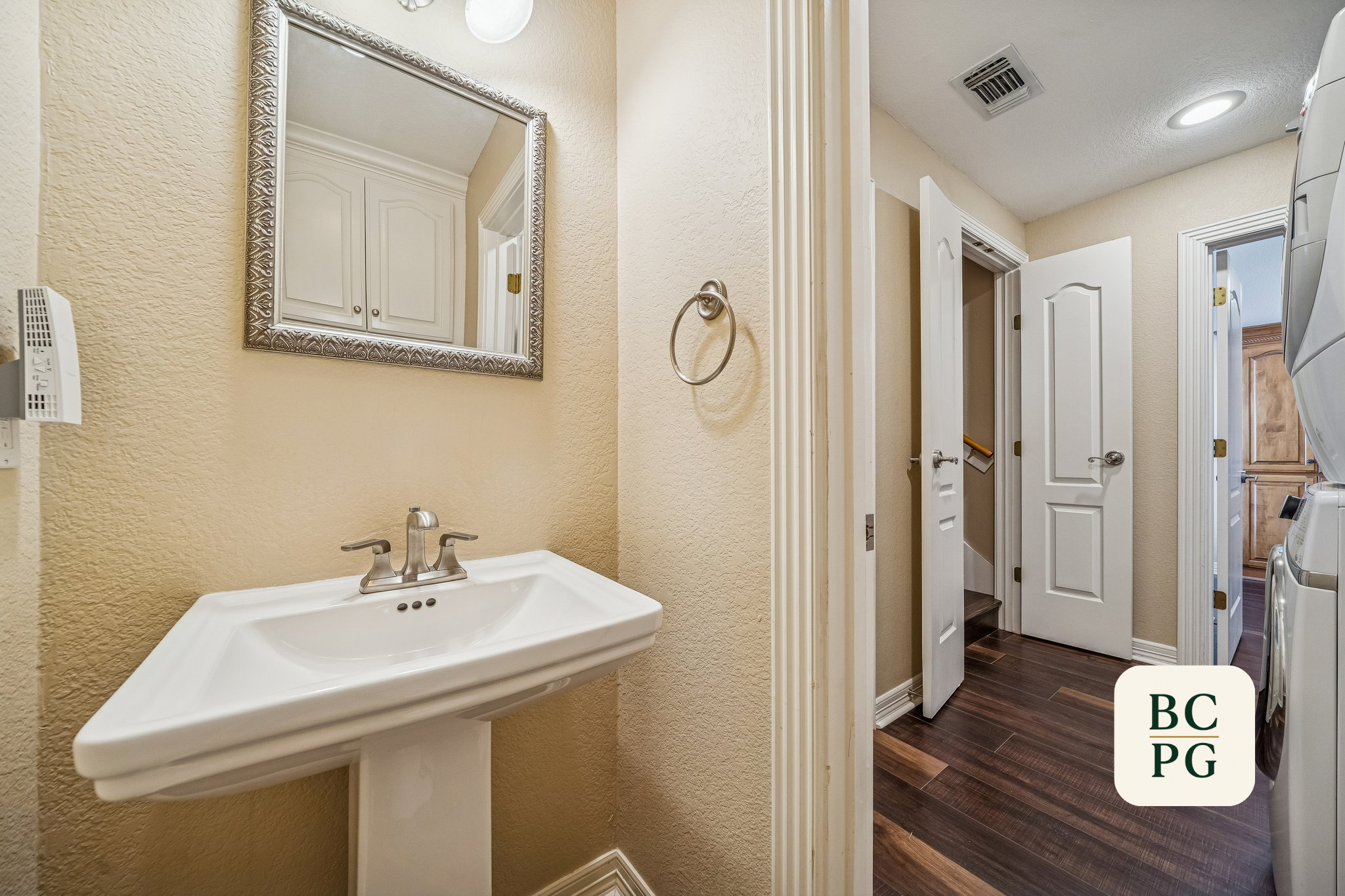 View of a small bathroom sink with a faucet, mirror, and beige textured wall. Adjacent is a laundry area with washing machines and open doors, hardwood flooring, and a ceiling vent.
