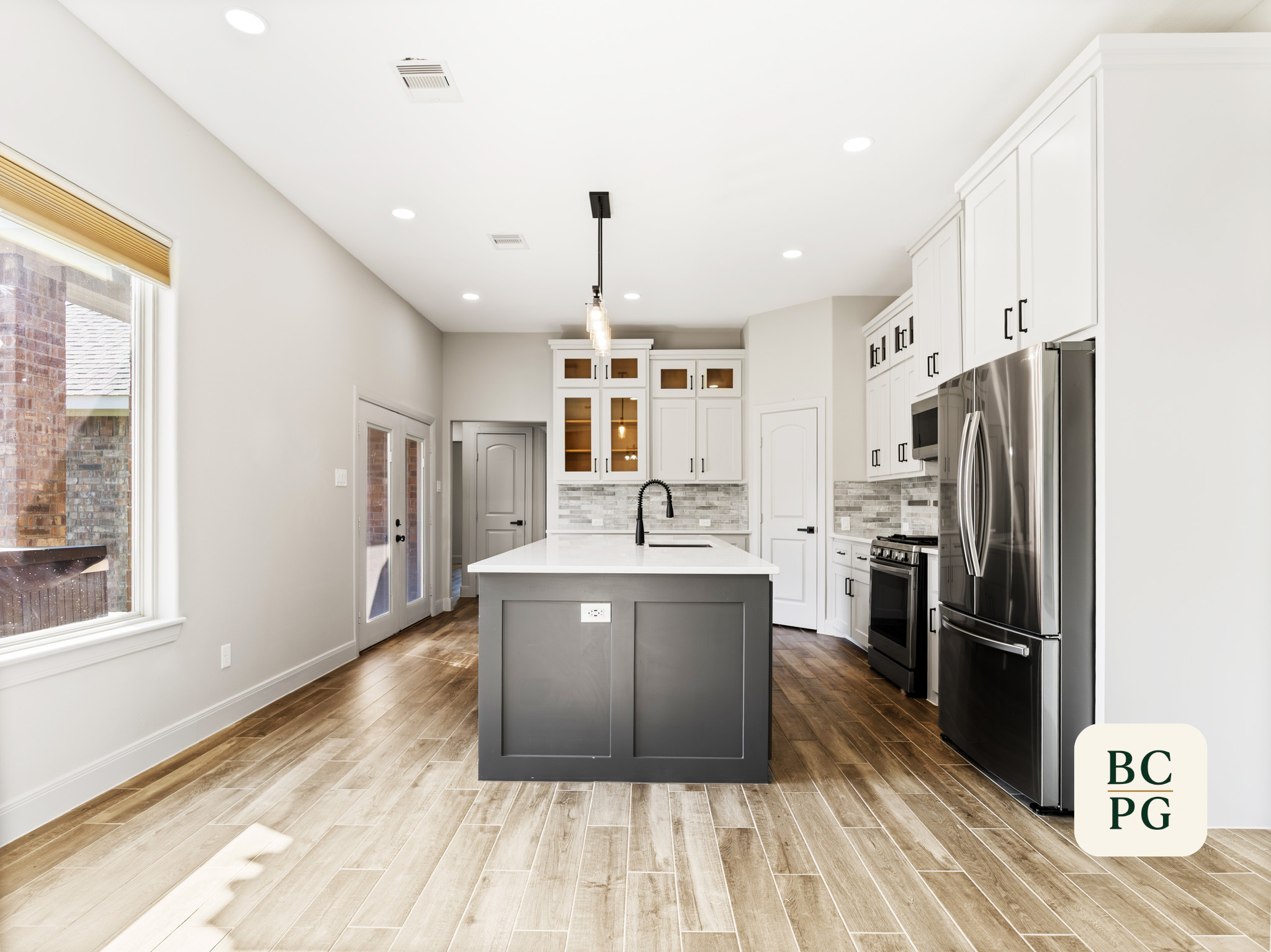 Modern kitchen with white cabinets, stainless steel refrigerator, black oven, white island with black faucet, wood flooring, large window, and white walls.