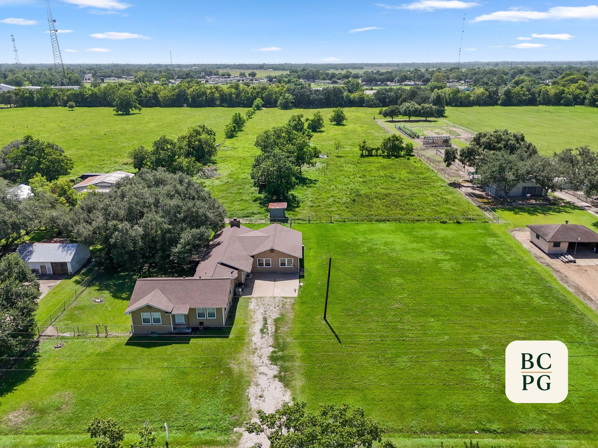 Aerial view of a rural residential area with houses, green lawns, and trees, surrounded by open fields and a distant cityscape with communication towers under a partly cloudy sky.