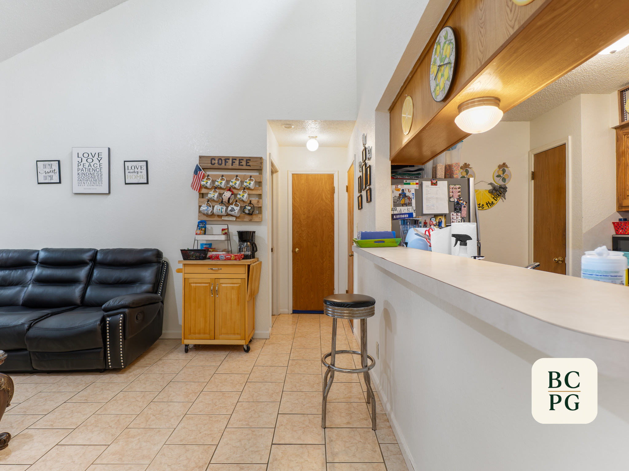 Interior of a home with a black leather sofa on the left, a kitchen counter with barstools in the foreground, white walls decorated with framed art and a wooden mug rack with mugs, a coffee sign, and various household items.