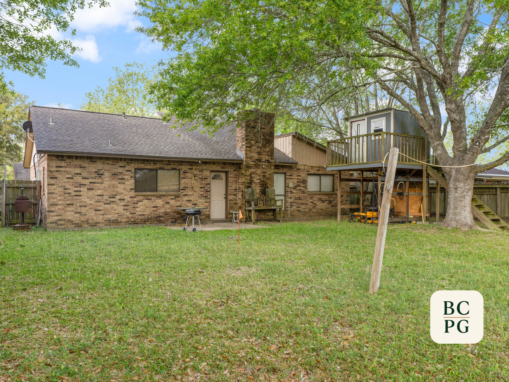 Backyard with brick house, large green tree, wooden deck, grass lawn, and various outdoor items.