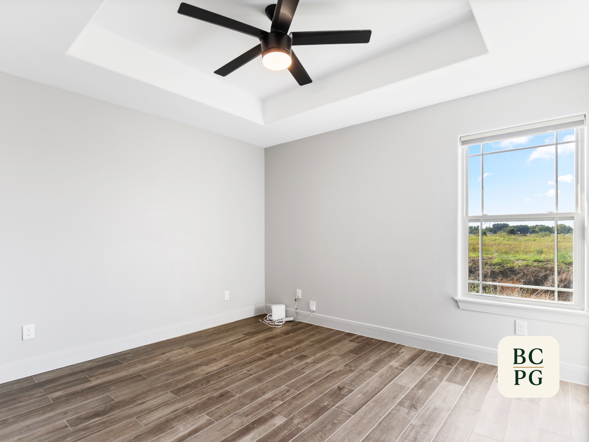 Empty room with white walls, wood flooring, a large window showing a grassy landscape, a ceiling fan with built-in light, and some electrical cords on the floor.
