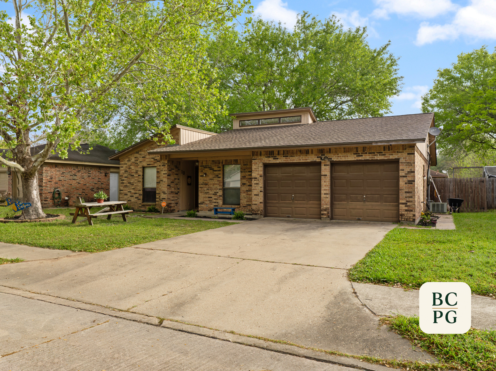 Front view of brick house with brown garage doors, concrete driveway, and attached porch, surrounded by green lawn, large trees, and a blue sky with clouds.