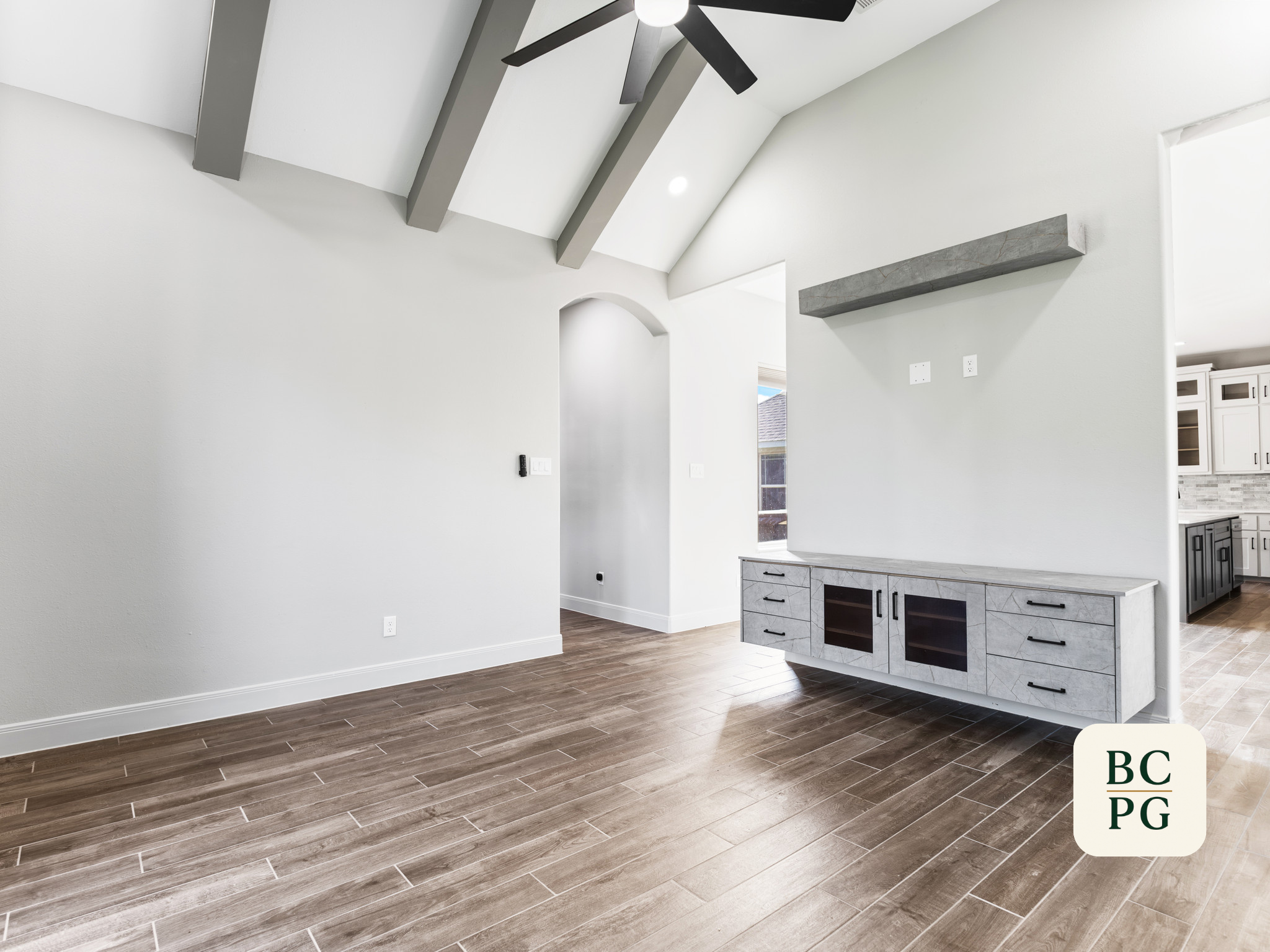 Interior of a modern home with white walls, wood-look floor tiles, a ceiling fan, and a kitchen area with grey cabinets and a white countertop.