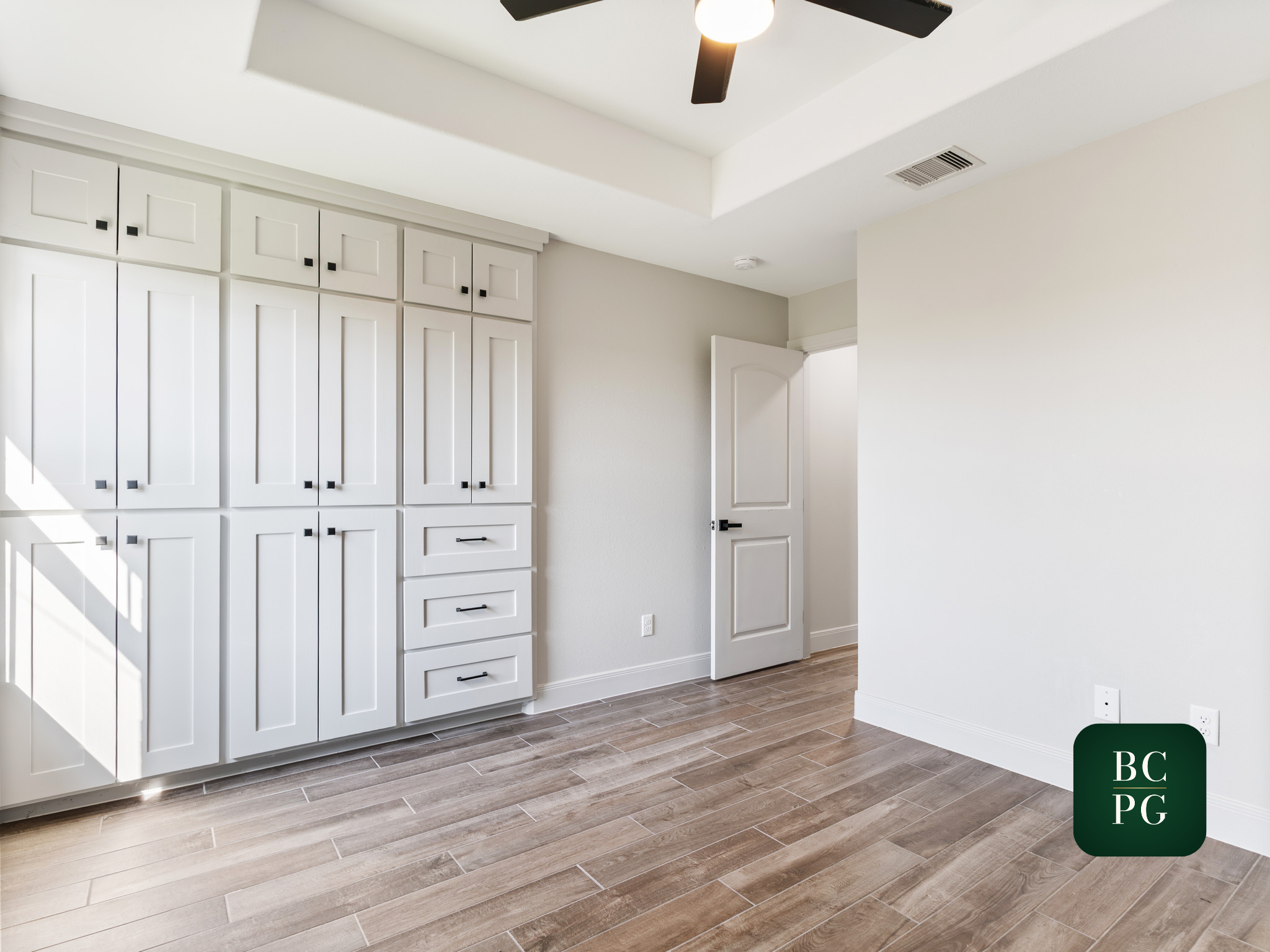A room with white cabinetry on the left side, an open door on the right side, a ceiling fan, and wood-look tile flooring.