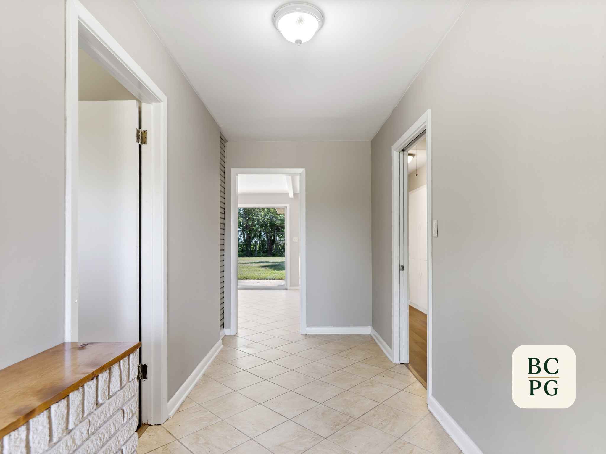 Interior hallway with white walls and tile floor, doorways on both sides, open to a sunlit outdoor area with trees.