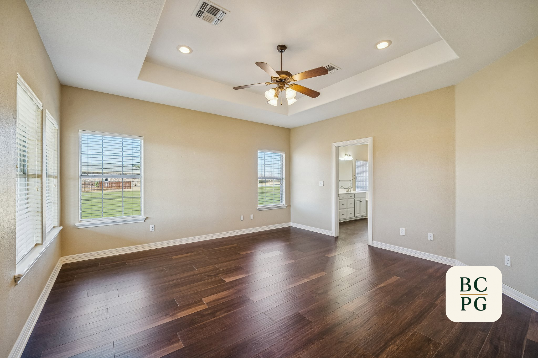 Empty living room with beige walls, dark wood flooring, multiple windows with blinds, a ceiling fan with lights, and a doorway leading to a bathroom.