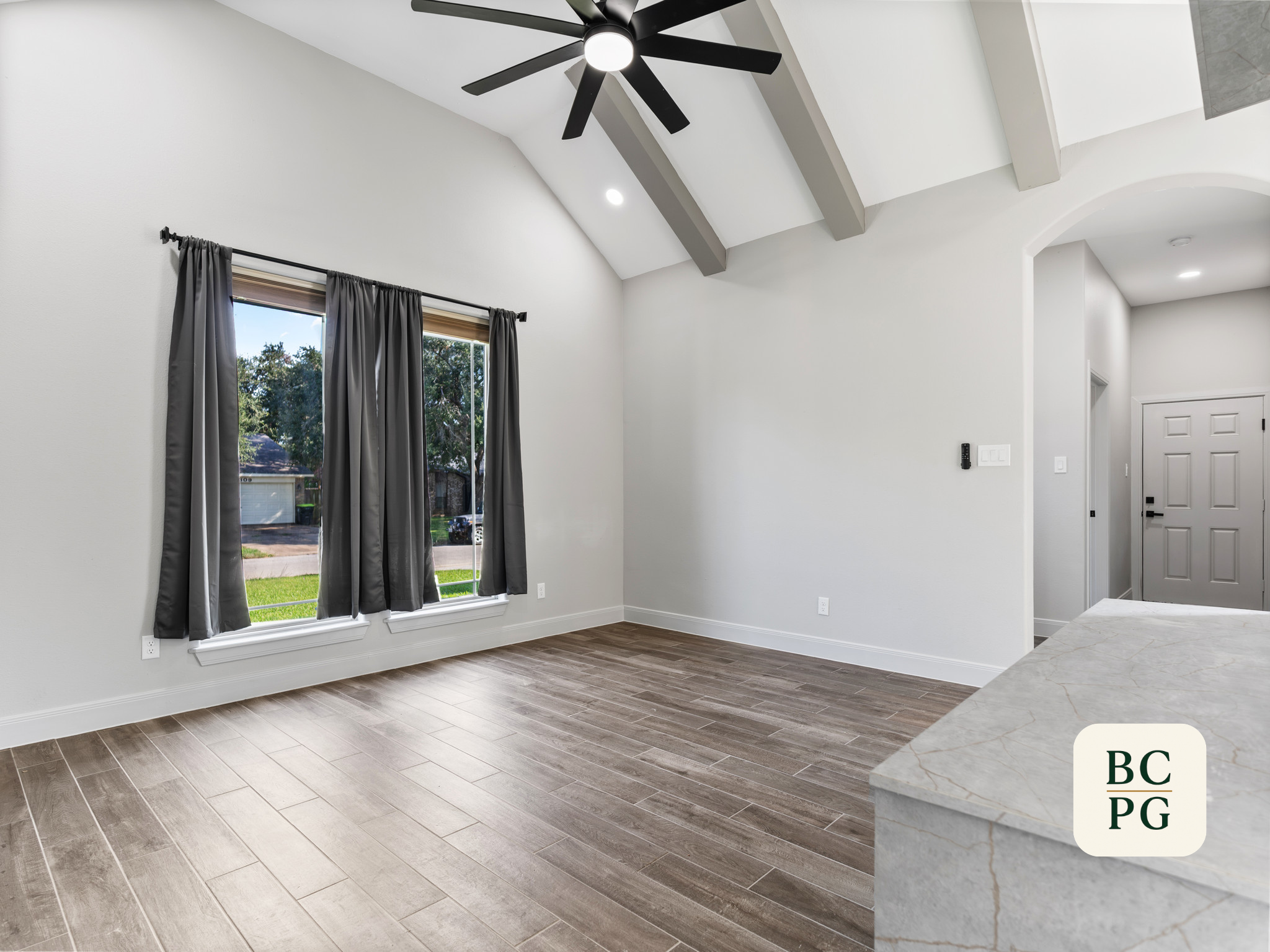 Interior of a modern living room with vaulted ceiling, black ceiling fan, two large windows with gray curtains, laminate wood flooring, and a white door in the corner.