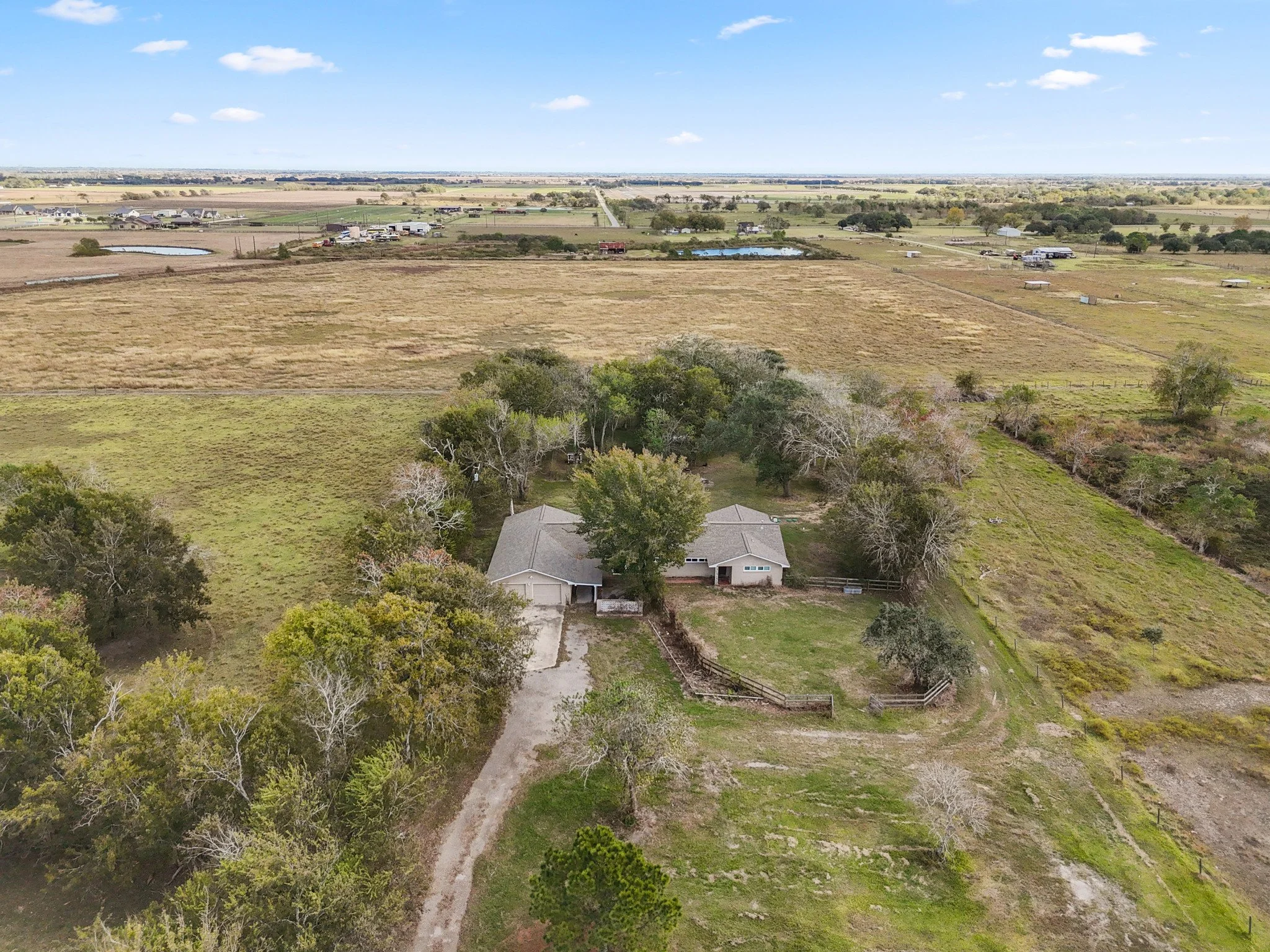 An aerial view of a rural property featuring a single-story house surrounded by trees, open grassy areas, and fenced-in sections, with expansive farmland and fields visible in the background under a partly cloudy sky.