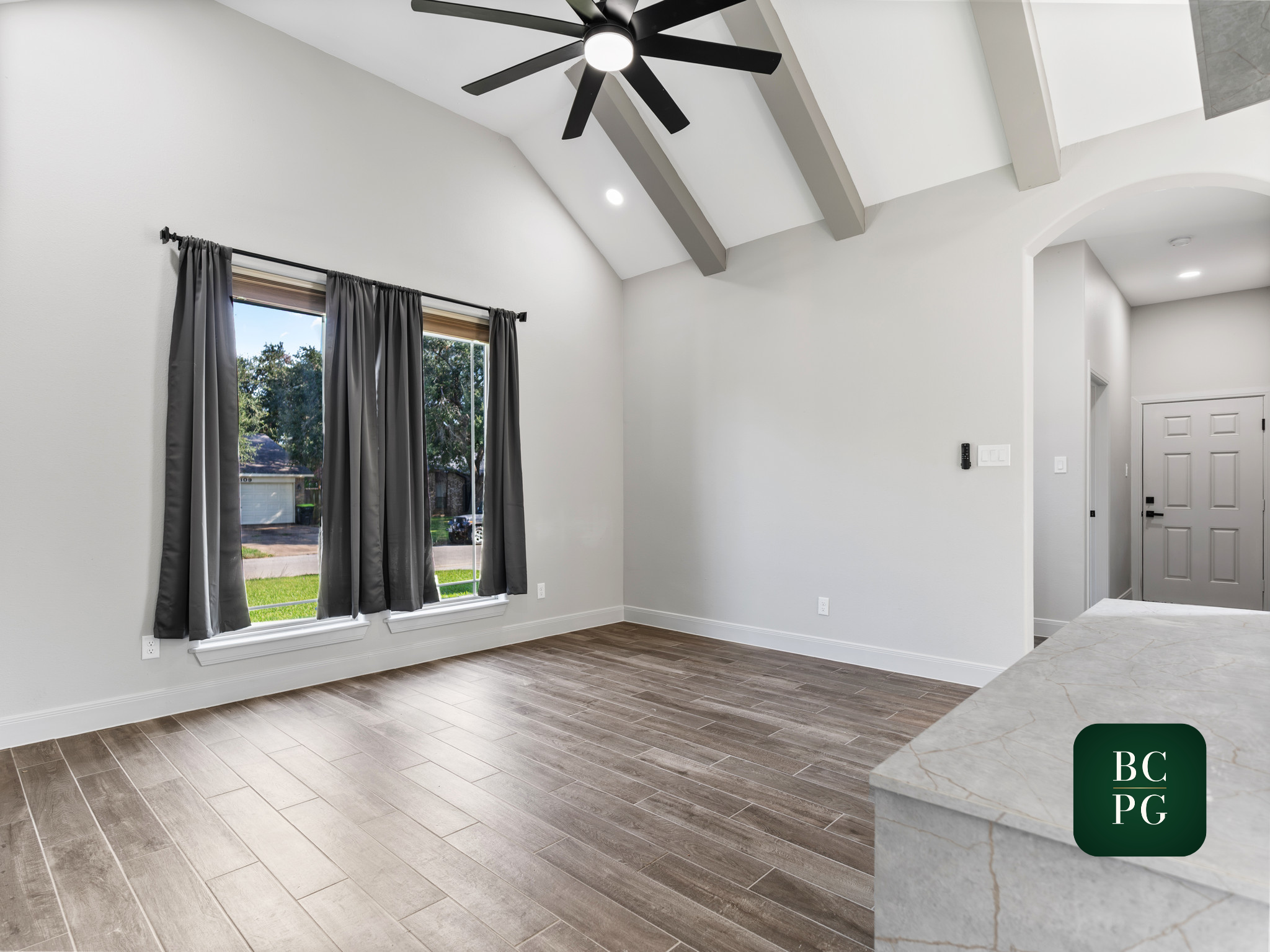 Interior view of a modern kitchen and living area with white walls, a vaulted ceiling with gray beams, a ceiling fan with black blades, two large windows with gray curtains, and hardwood flooring.