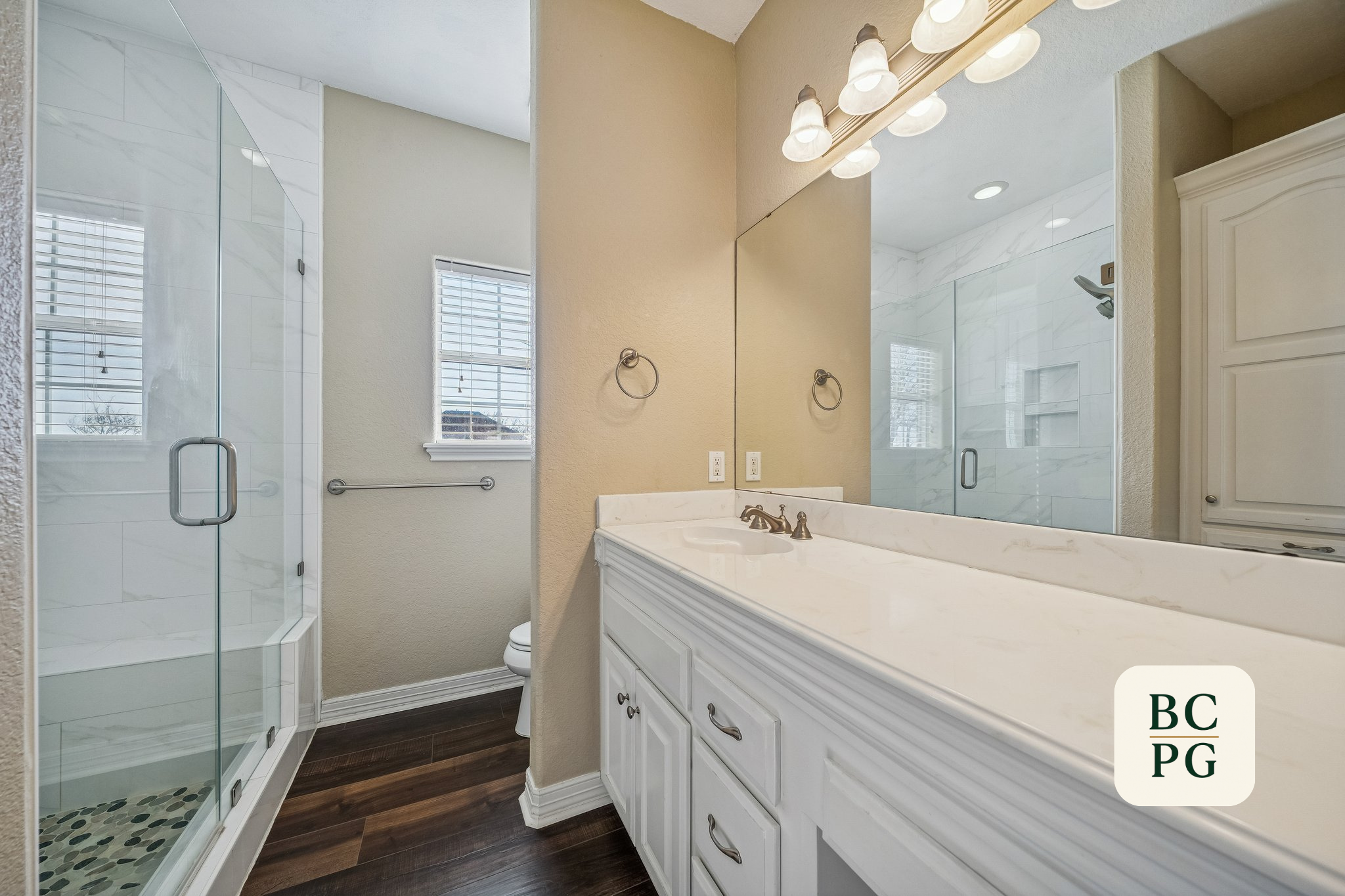 A bathroom featuring a glass-enclosed shower on the left, a white vanity with a marble countertop and sink, and a large mirror above it. The bathroom has beige walls, two green towels, and two windows with blinds.
