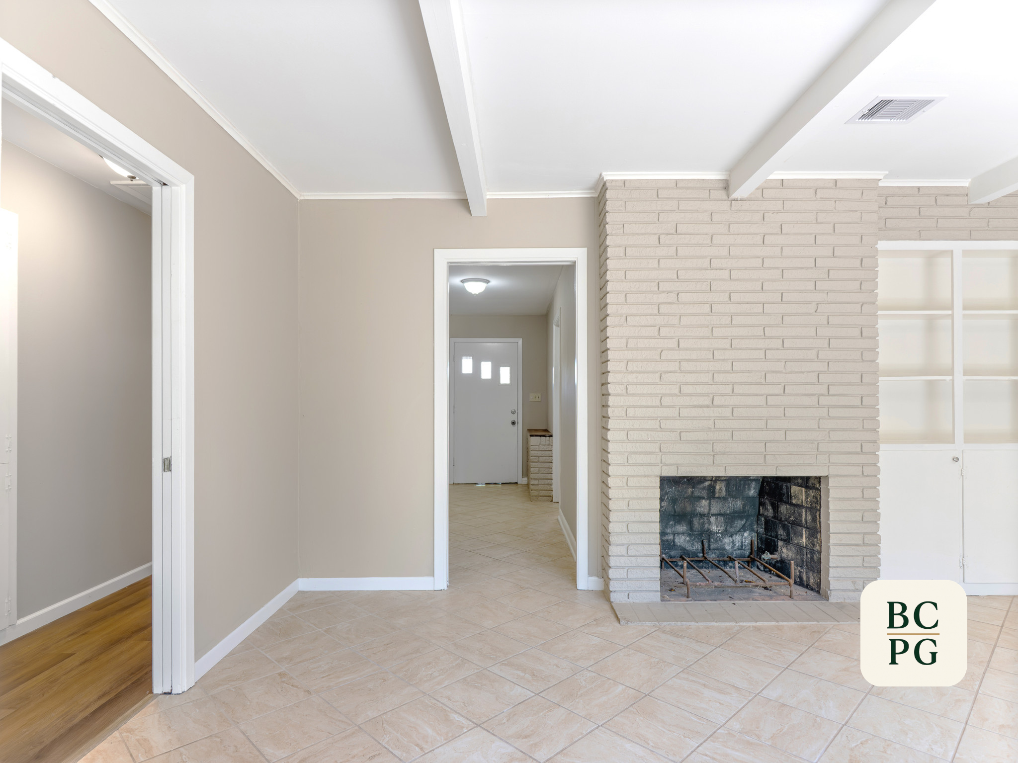 Interior of a house with beige walls, white trim, a brick fireplace, and tiled flooring. Doorways lead to other rooms, and a foyer with a front door is visible in the background.
