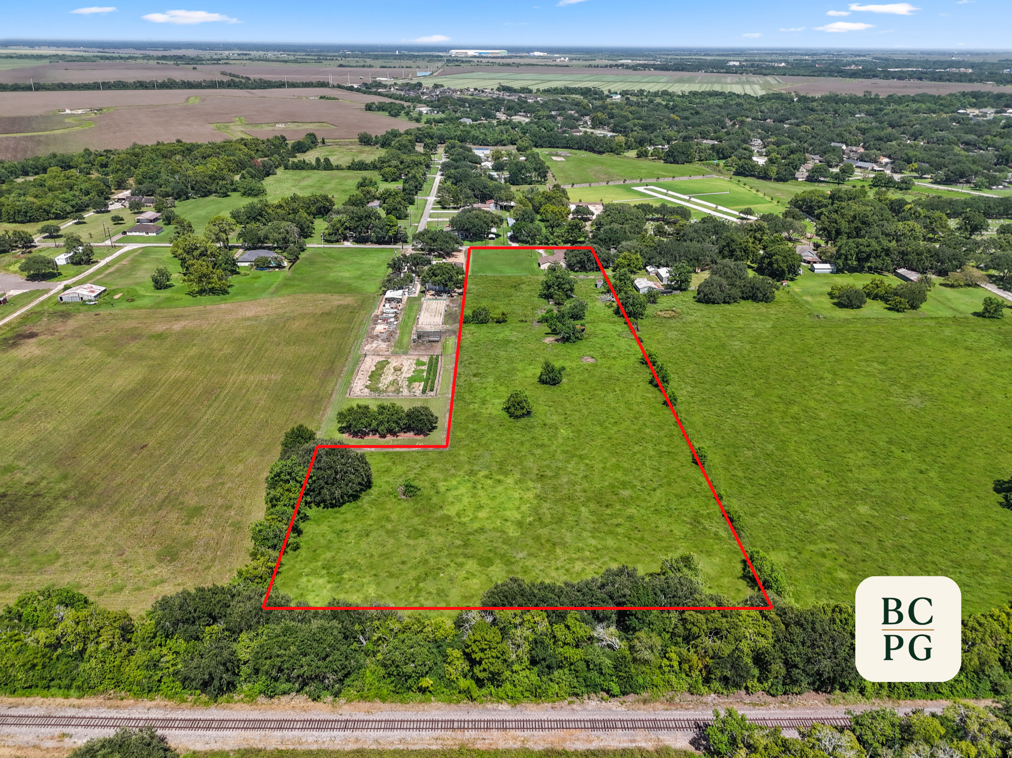 Aerial view of a rural area with green fields, trees, and houses, with a red boundary line outlining a specific plot of land. The background shows more farmland and a few distant buildings.