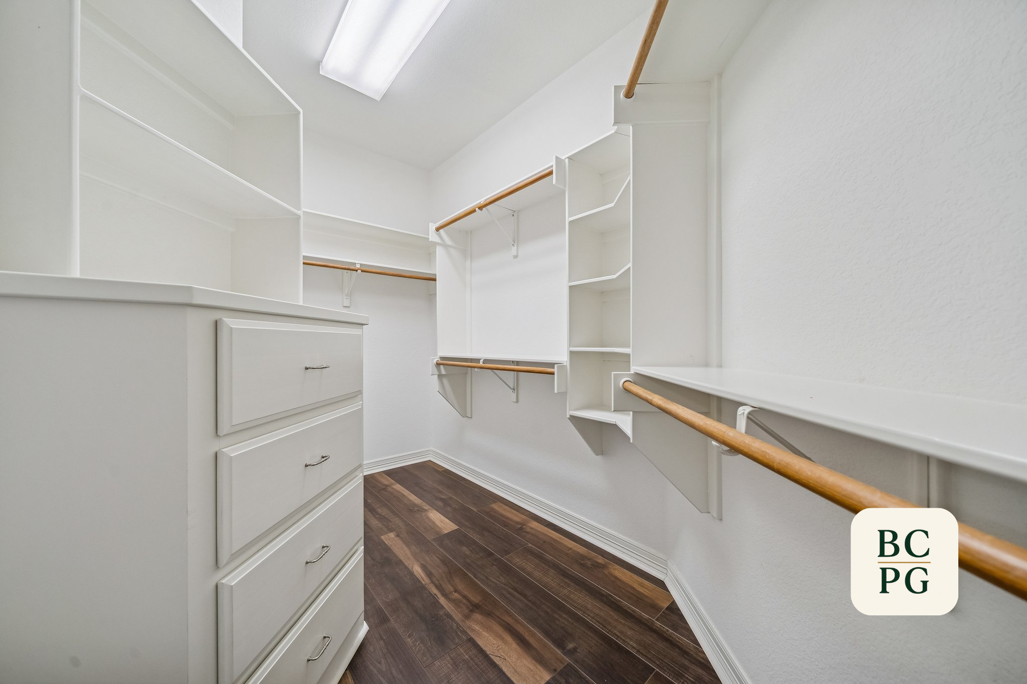 Empty walk-in closet with white shelving units, wooden closet rods, and dark wooden flooring.