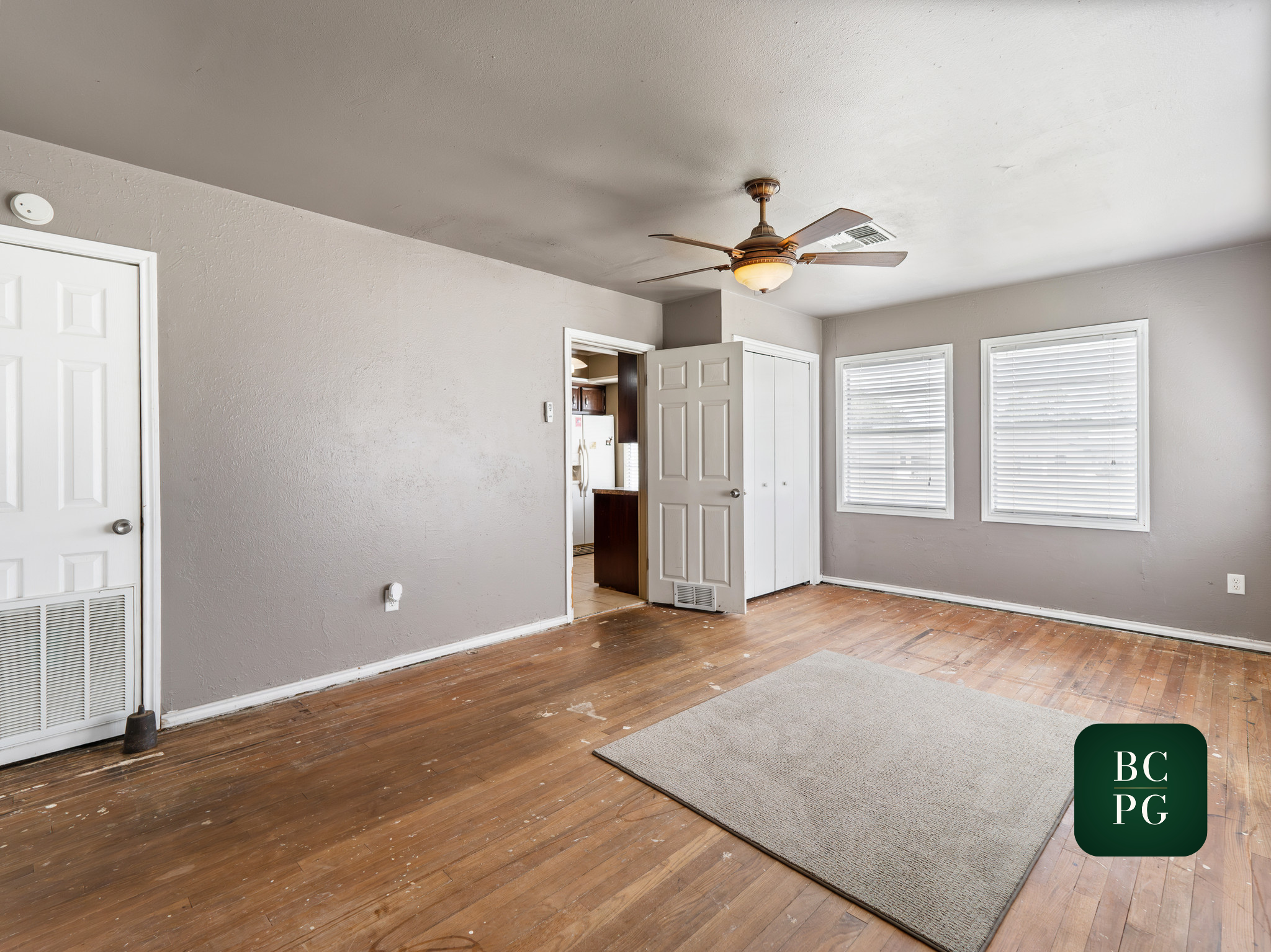 Interior of a room with light gray walls, wooden flooring, a ceiling fan, multiple windows with white blinds, a partially open door leading to a kitchen area, and a small section of a carpeted floor in the foreground.