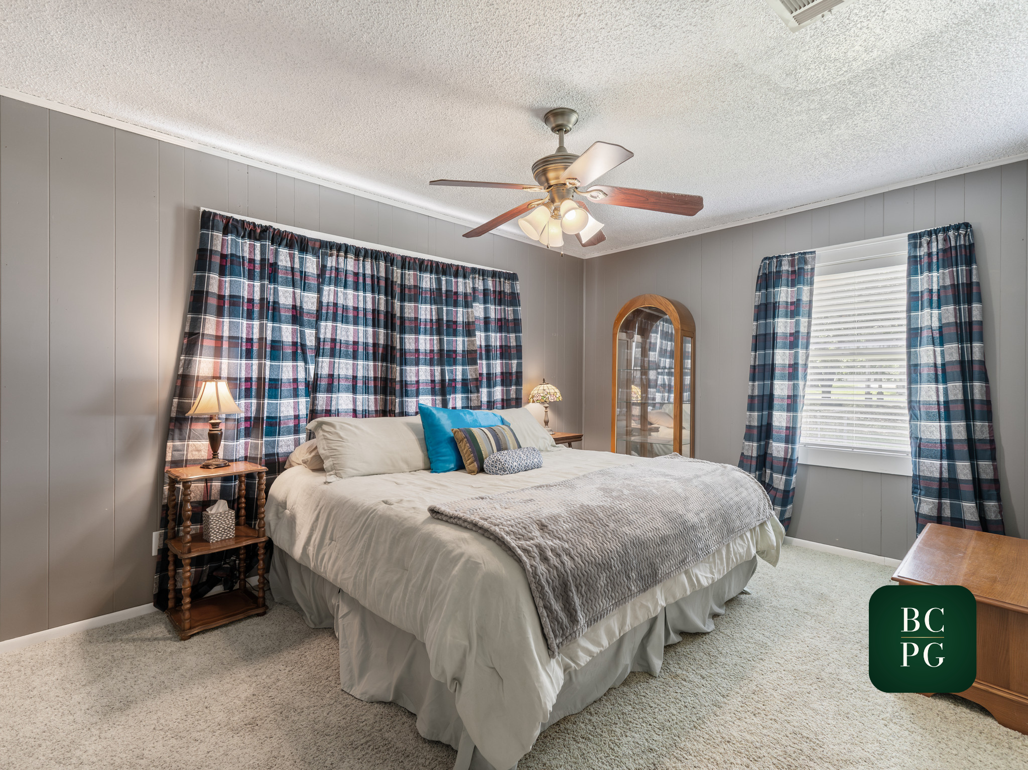 A bedroom with gray paneled walls, a bed with white bedding and colorful pillows, checkered curtains, a wooden nightstand with a lamp, and a ceiling fan with lights.