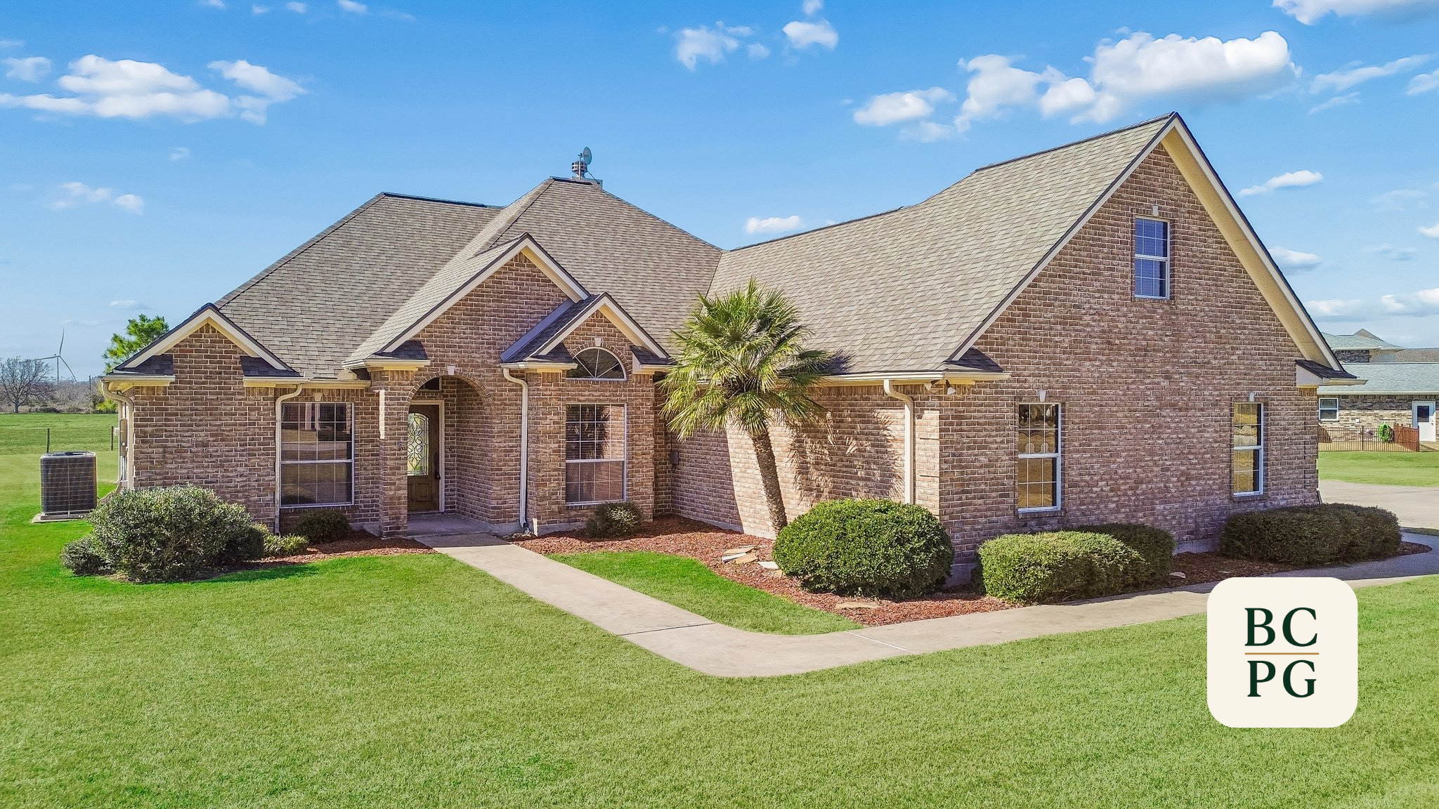 A two-story brick house with a gabled roof, front porch, and multiple windows. The house is surrounded by a well-maintained lawn with bushes and a small palm tree. The sky is bright and partly cloudy.