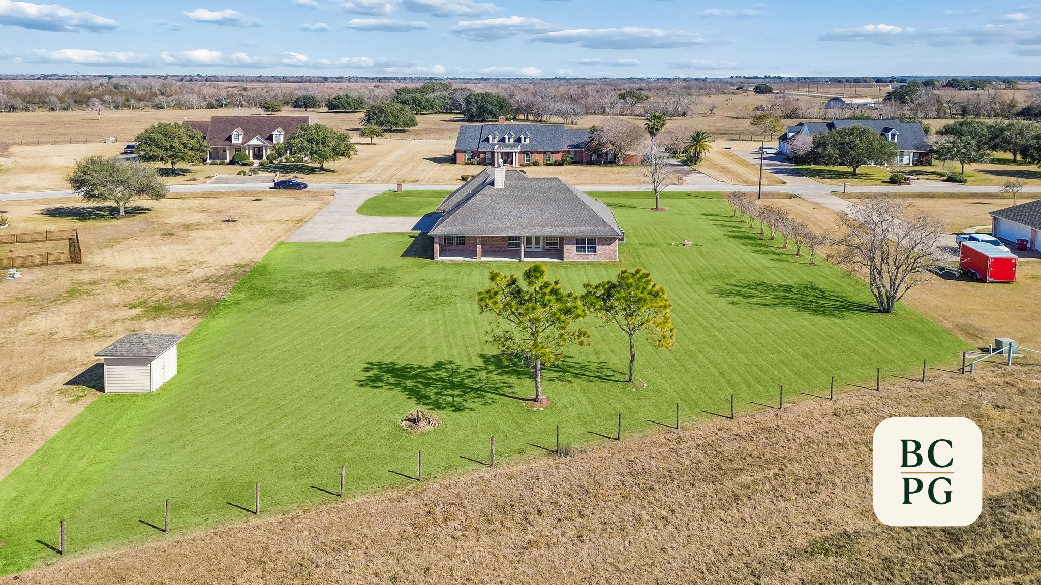 Aerial view of a suburban neighborhood with houses, trees, and well-maintained yards, including a house with a large green lawn and a separate small shed, under a partly cloudy sky.