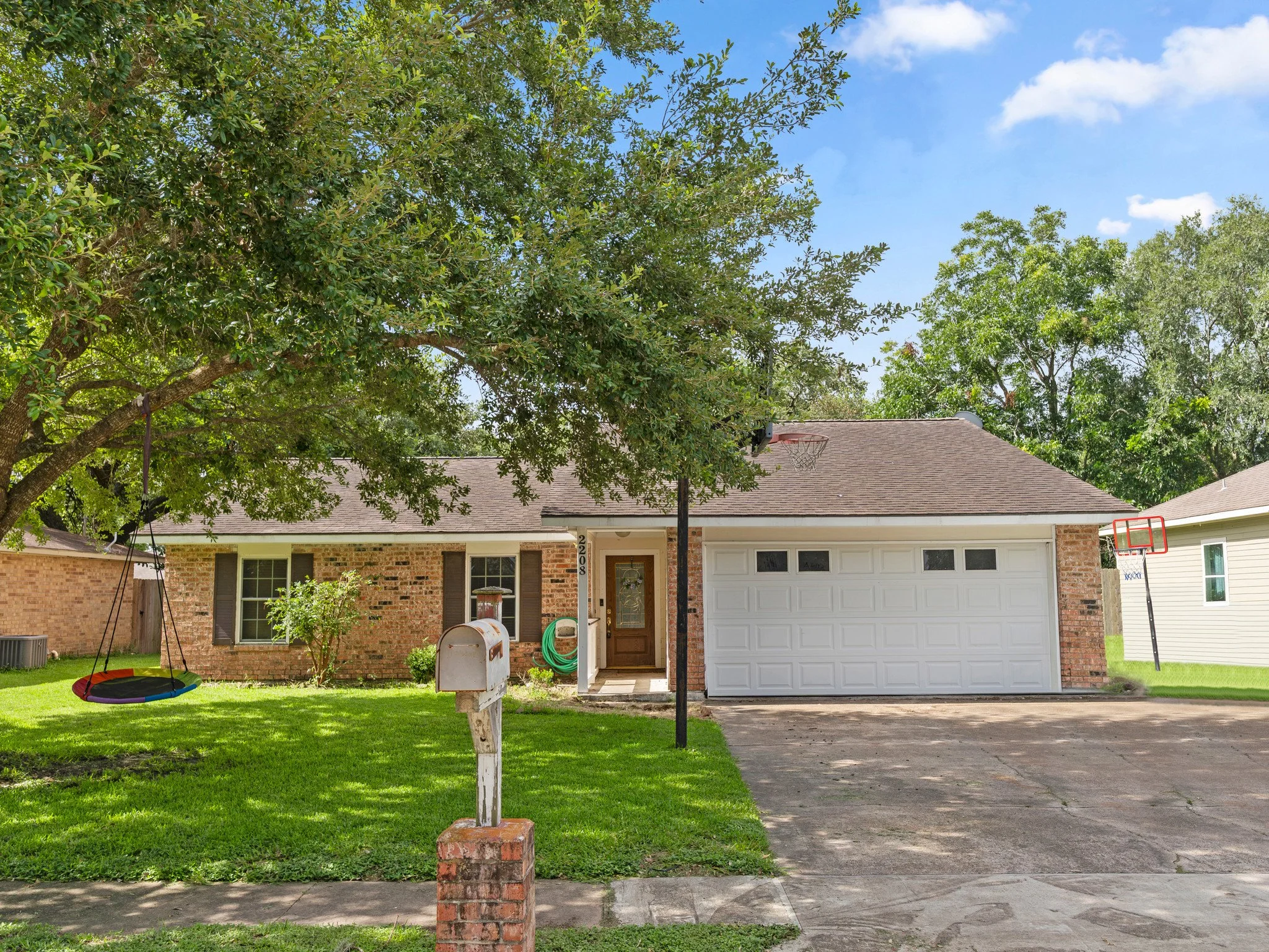 A suburban house with a brick exterior and attached garage, front yard with green lawn, tree, swing, mailbox, and nearby basketball hoops, under a partly cloudy sky.