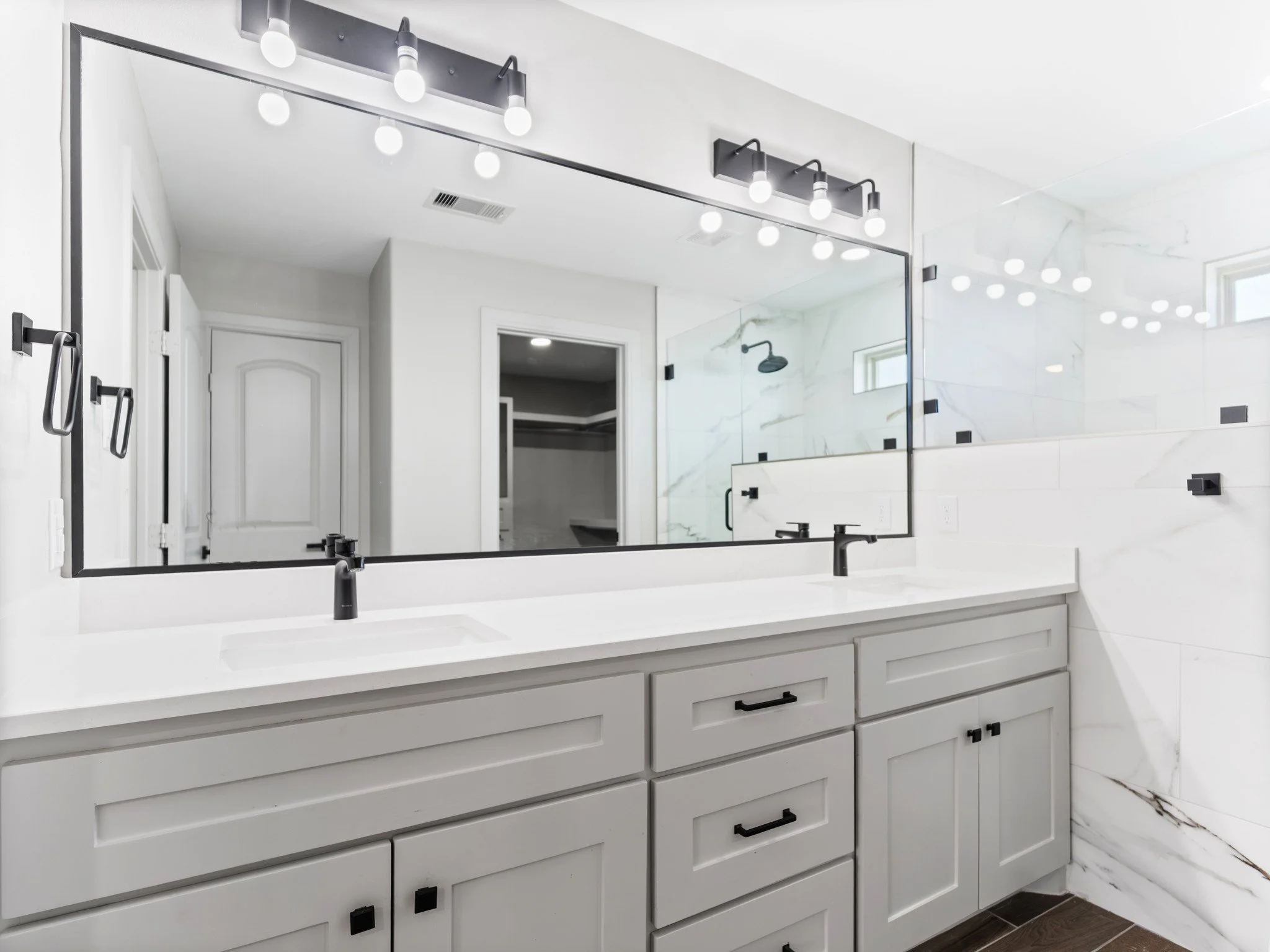 Modern bathroom with white double vanity, large mirror, black fixtures, marble tiles, and a walk-in shower in background.