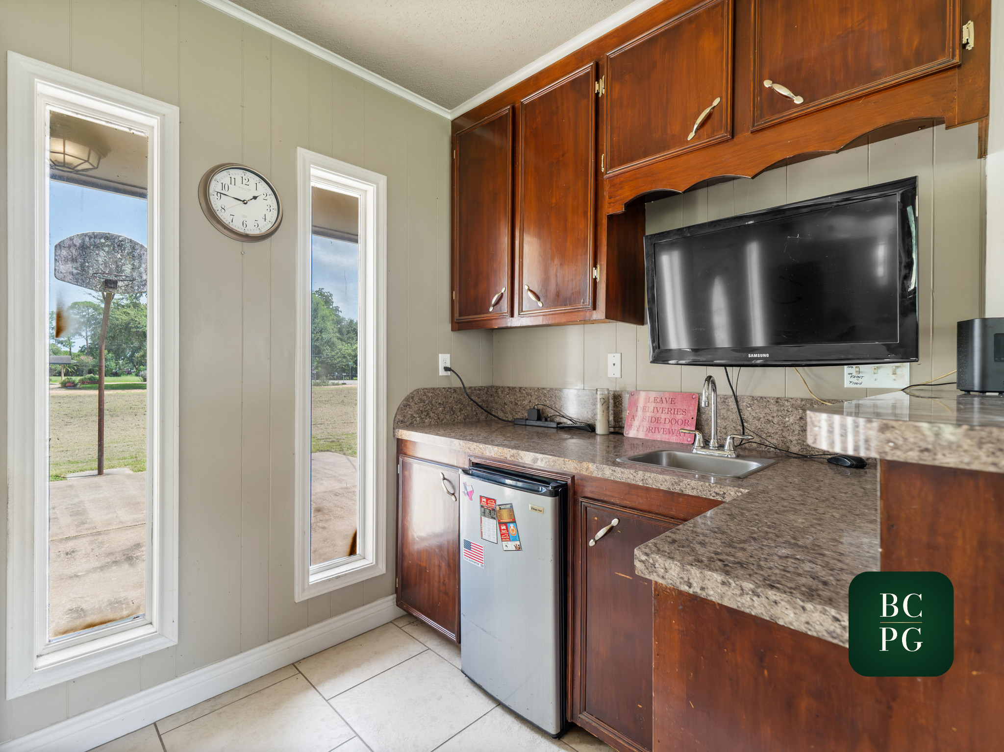 Kitchen corner with a textured beige tile floor, light green paneled wall, corner cabinets with a dark wood finish, a mounted flat-screen Samsung television, a small sink, and a window showing trees outside. A clock is on the wall next to the window.