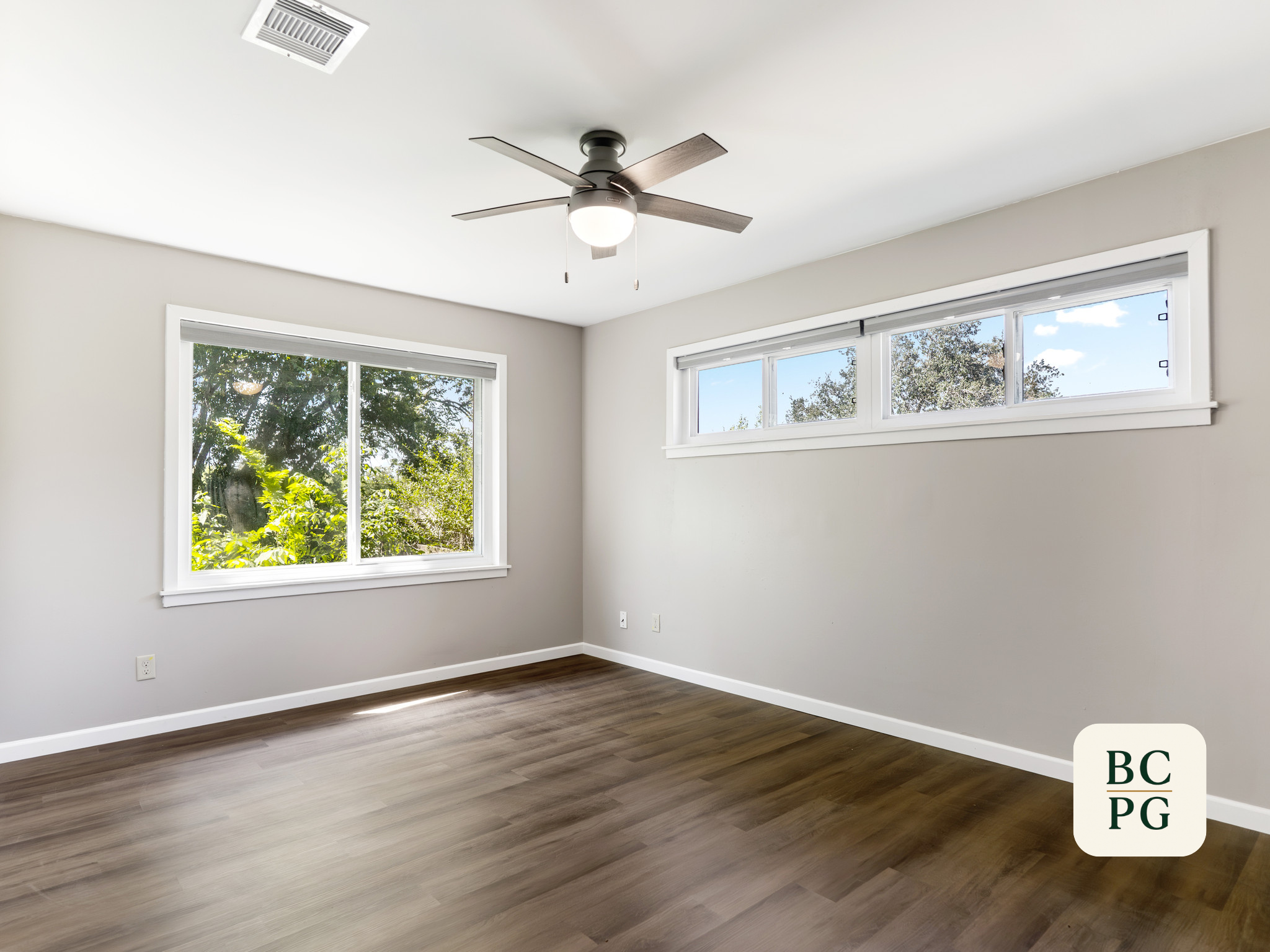 Empty room with large windows showing trees outside, white walls, dark wood flooring, and a ceiling fan with light.