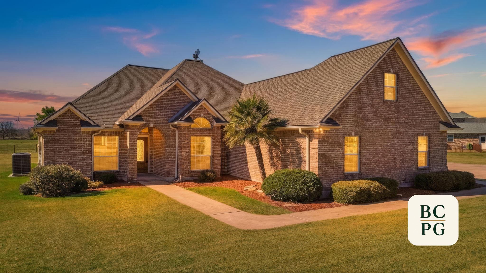 A brick house during sunset with a manicured lawn, bushes, a small tree, and windows glowing with interior light.