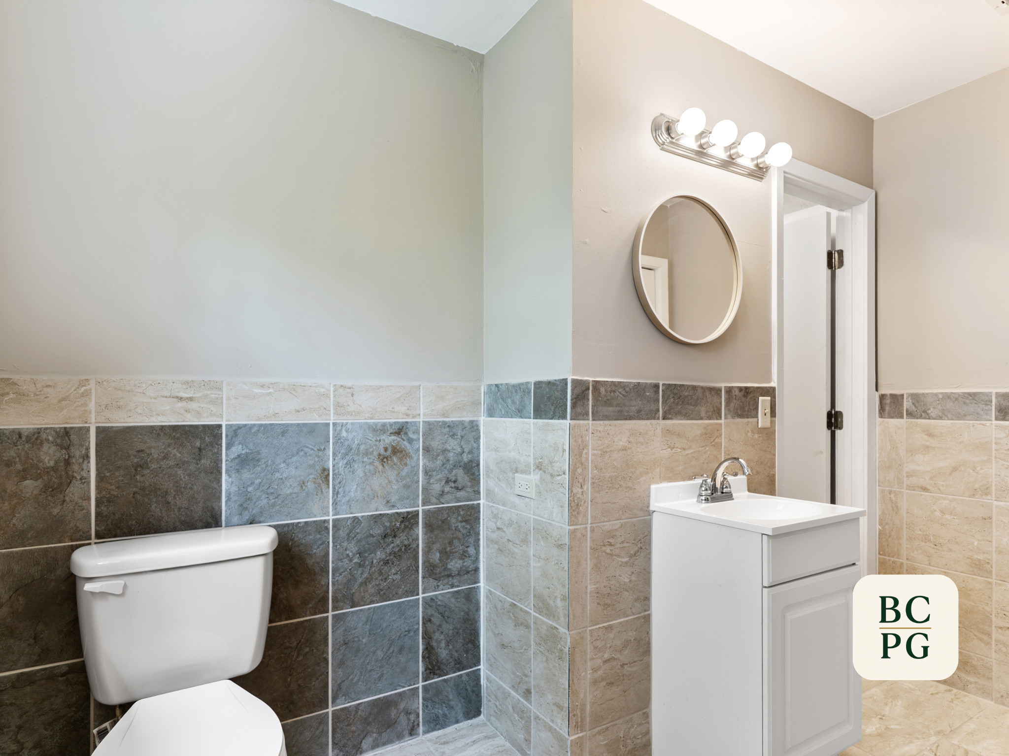 A bathroom with tiled walls, a toilet, a small sink with a cabinet, a mirror, and a light fixture with four bulbs above the mirror.