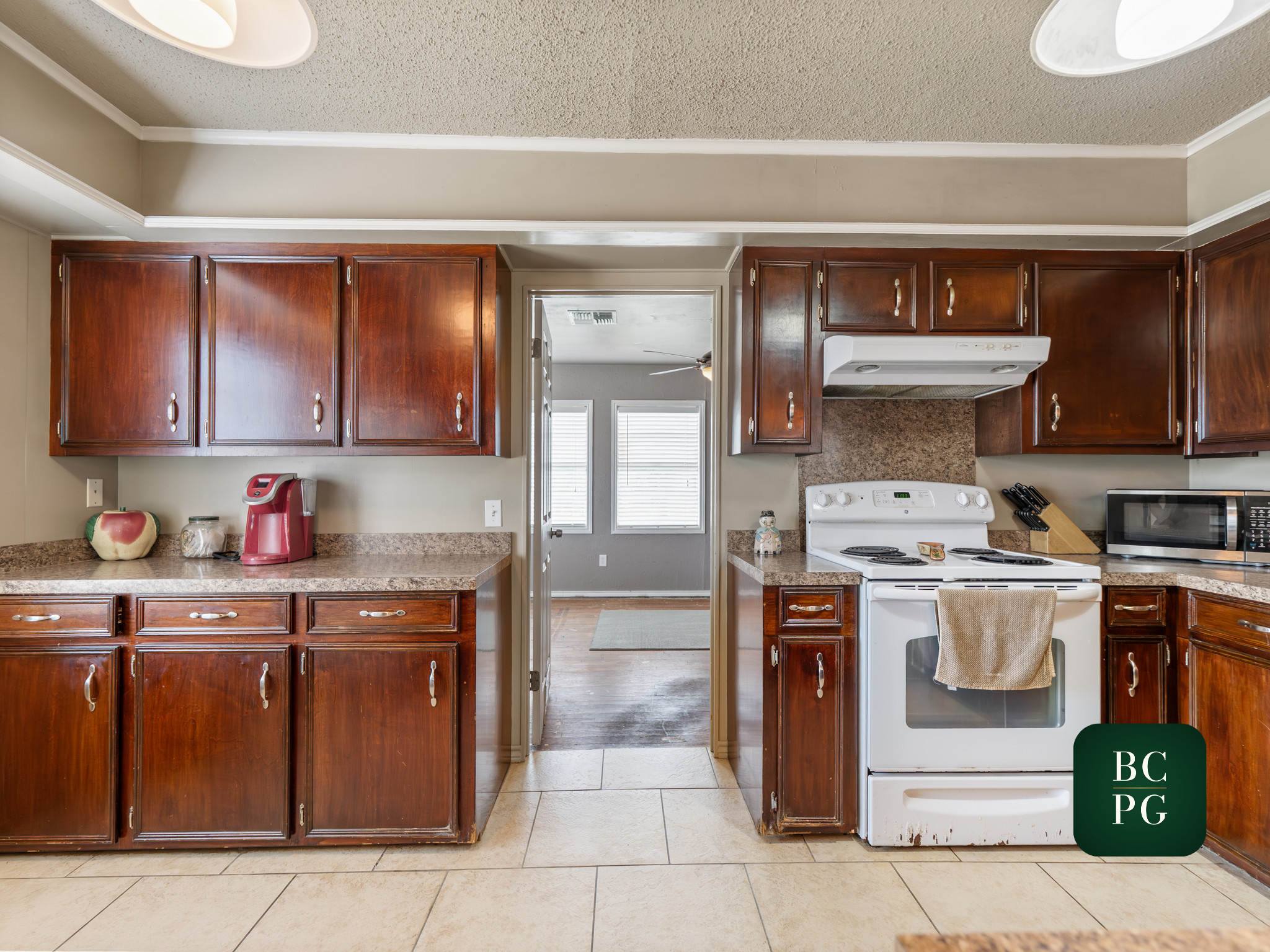 A kitchen with dark wooden cabinets, a white stove, a microwave, a red coffee maker, and a small decorative figurine on the countertop, with a doorway leading to a room with windows and a ceiling fan.