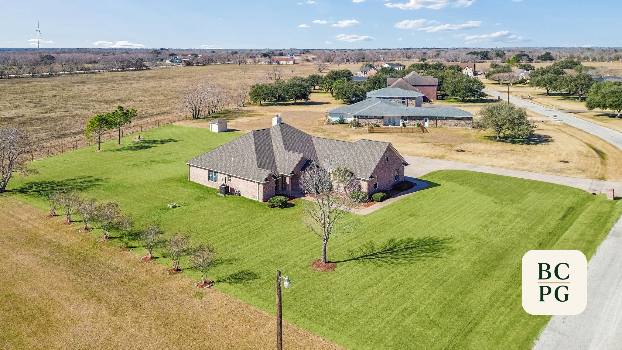 Aerial view of a suburban neighborhood with single-family houses, green lawns, trees, and a large open field in the background.