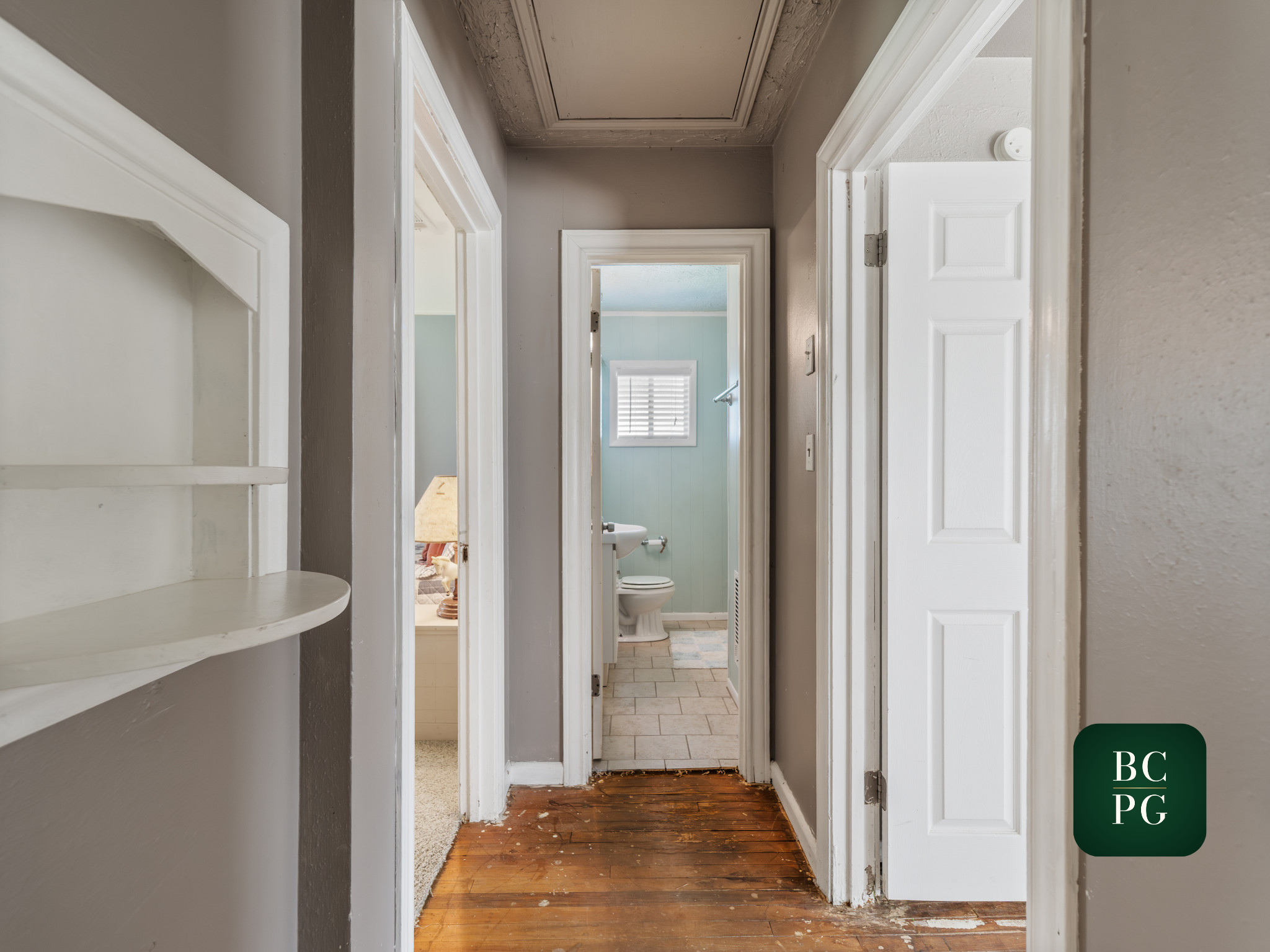 A view of a hallway with multiple doorways leading to different rooms, with a bathroom visible at the end. The flooring transitions from wooden to tile, and the walls are painted in light colors.