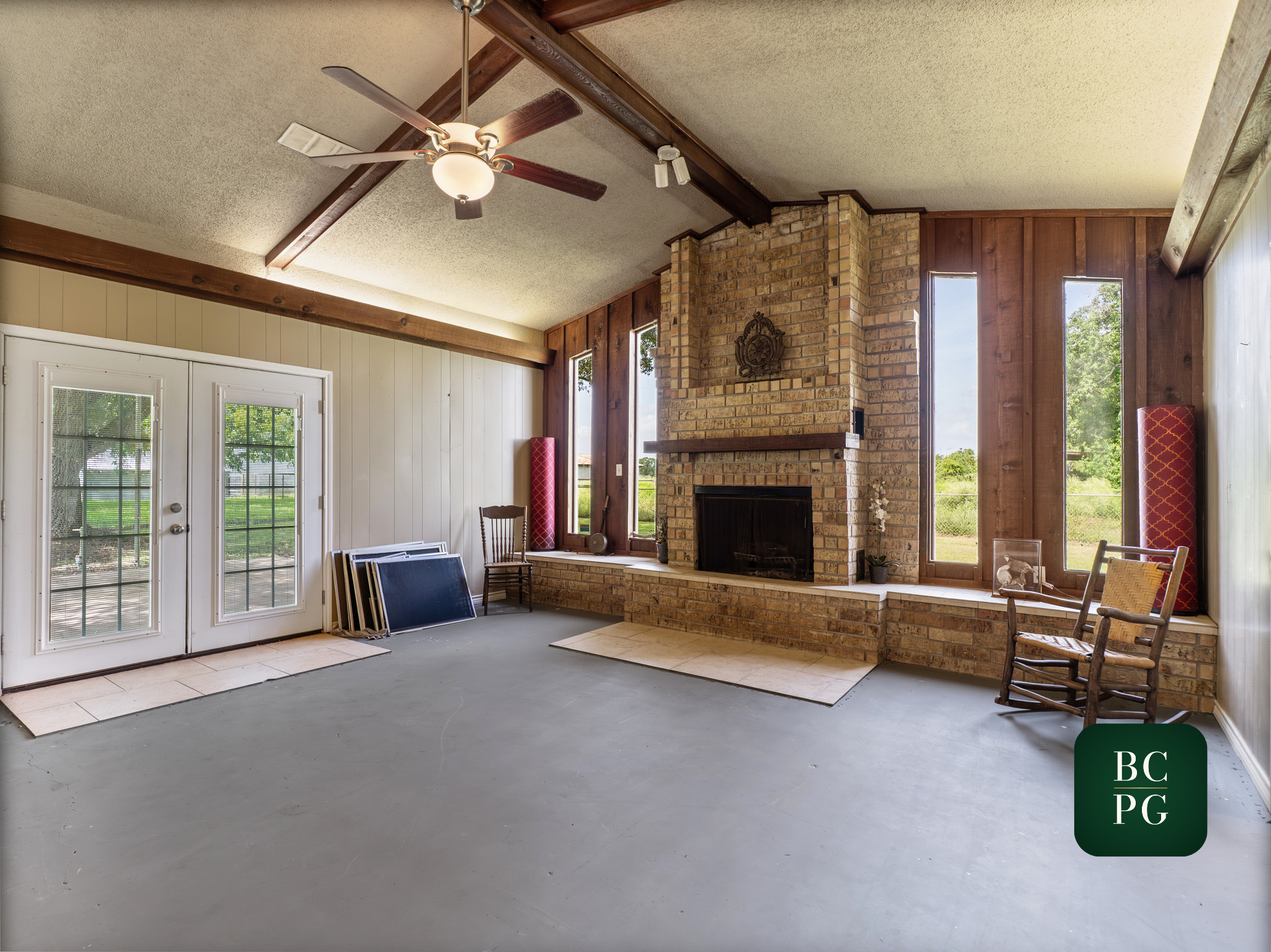 Living room with a brick fireplace, large windows, wooden wall panels, ceiling fan, and various chairs.